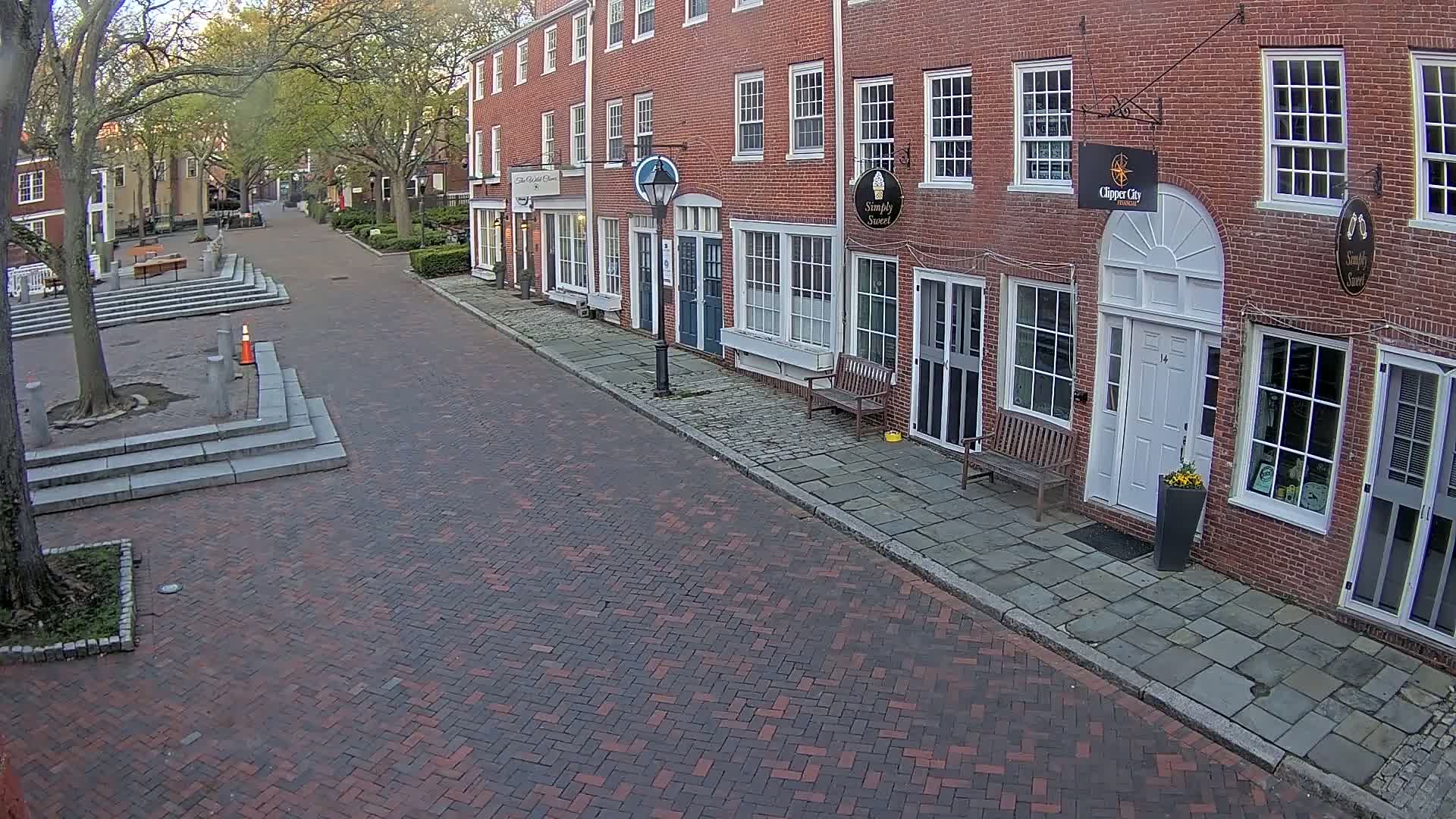 A brick-paved street lined with brick buildings on a partly cloudy day, with trees in full leaf along the street and a small plaza at the far end.