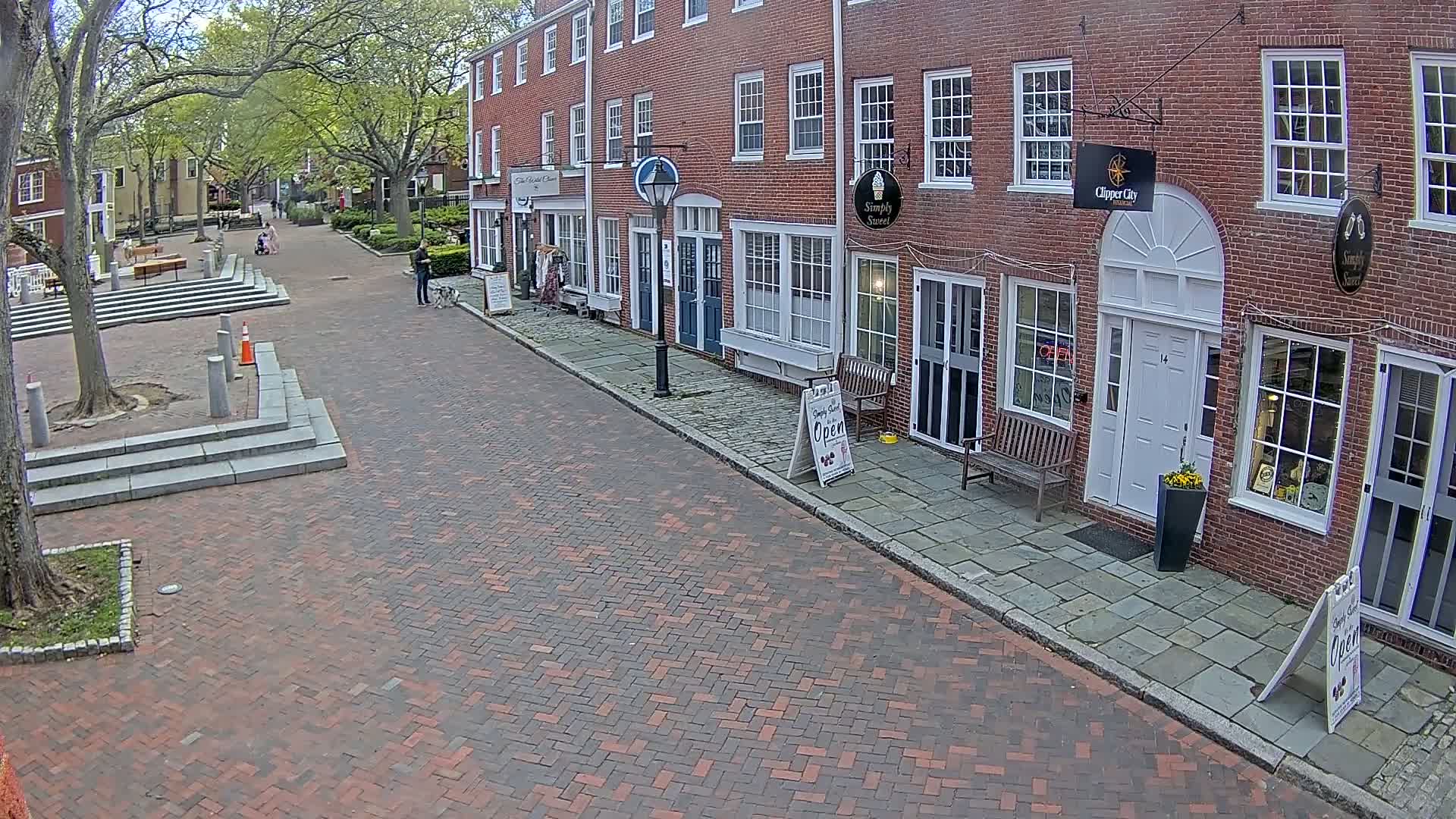 A brick-paved street lined with brick buildings under a partly cloudy sky, with a few pedestrians walking.
