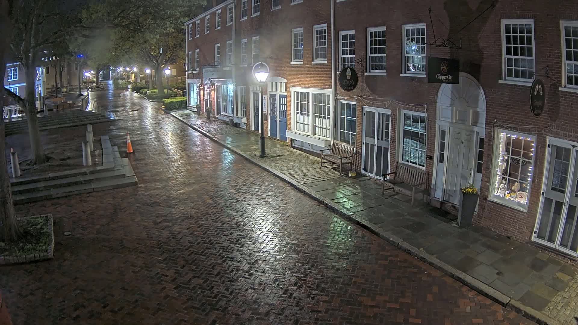 A rain-slicked brick street at night is lined with brick buildings, lit by streetlights and the soft glow from shop windows.