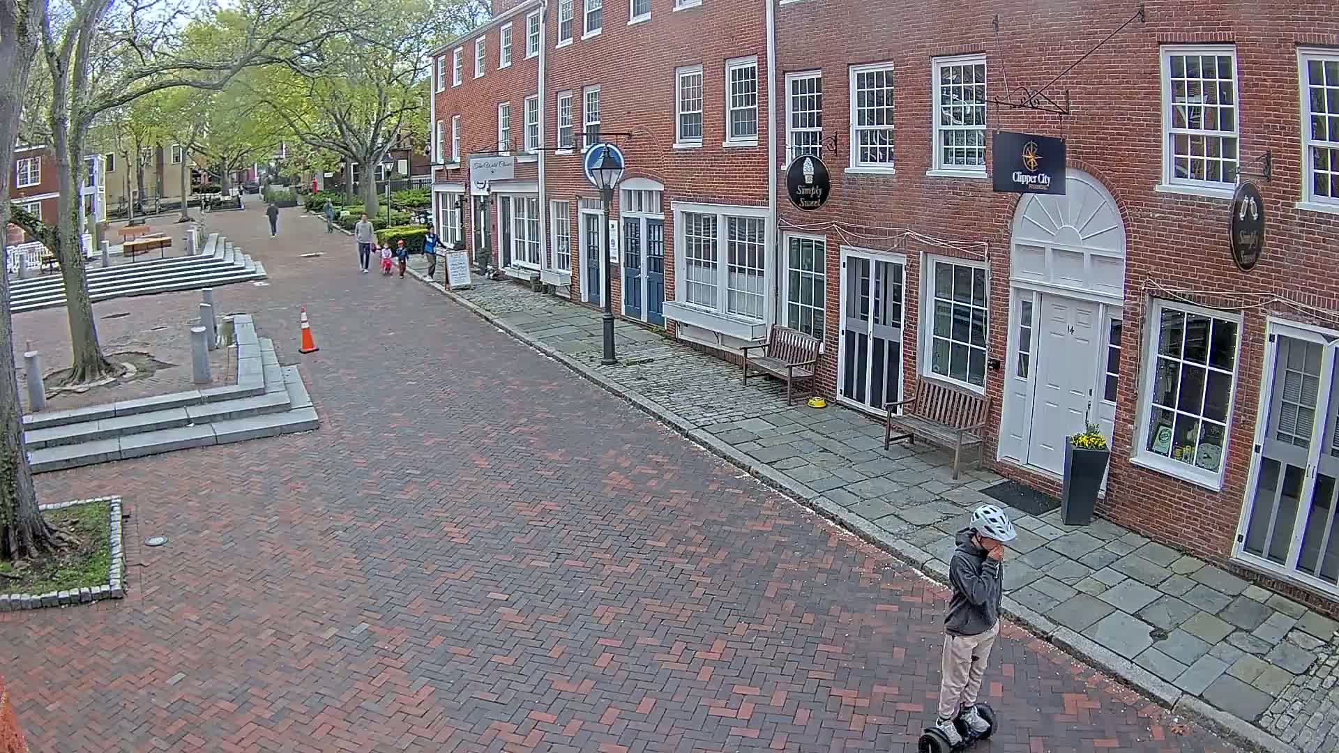 A person on a self-balancing scooter stands on a brick street in front of brick buildings on a partly sunny day.