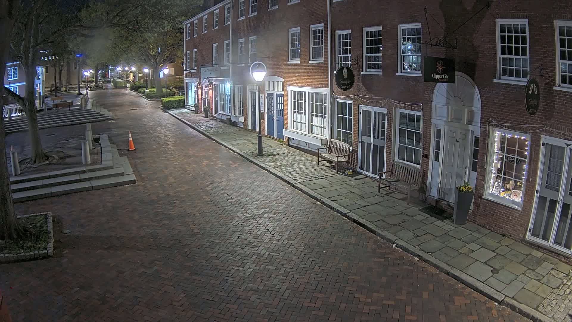 A brick street at night, lined with brick buildings, is lit by streetlights and displays a few potted plants.