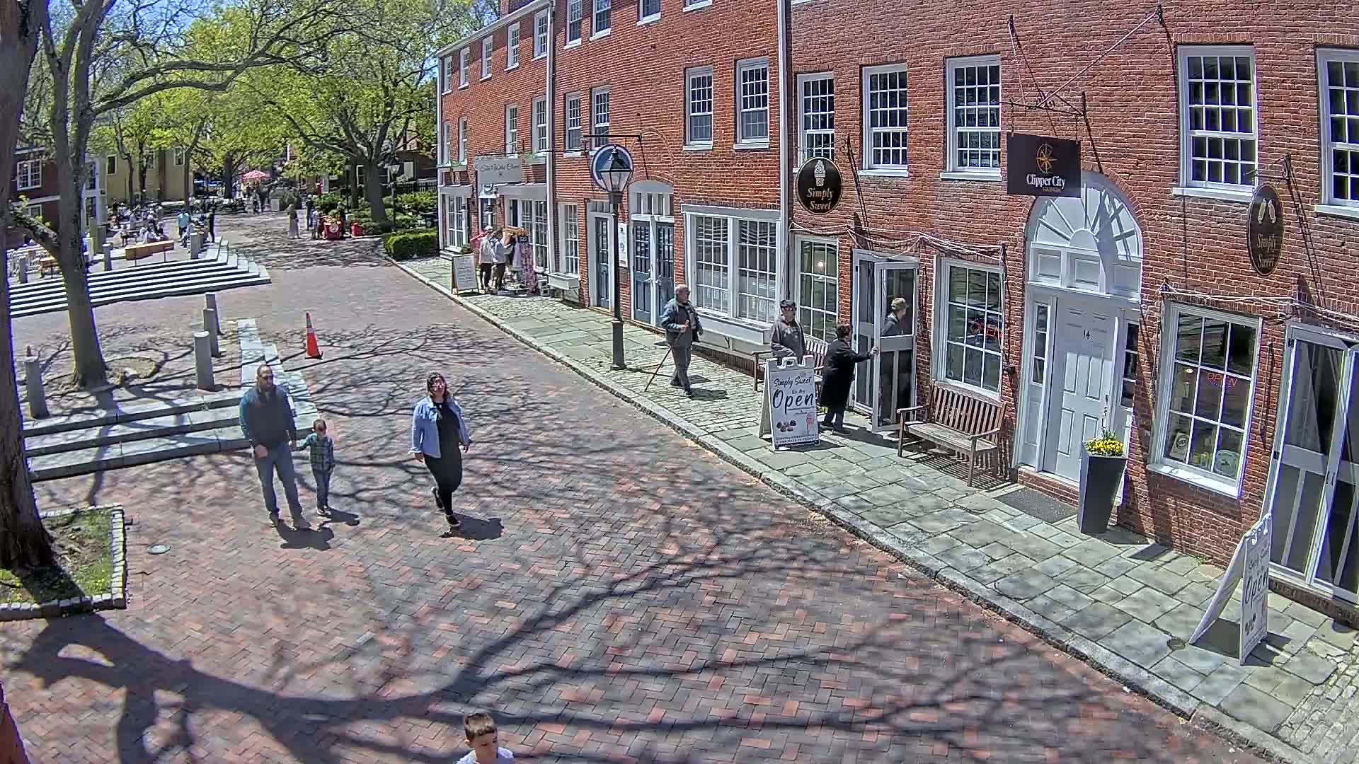 A brick street lined with brick buildings on a sunny day, with several people walking along the street and sitting in a nearby plaza.