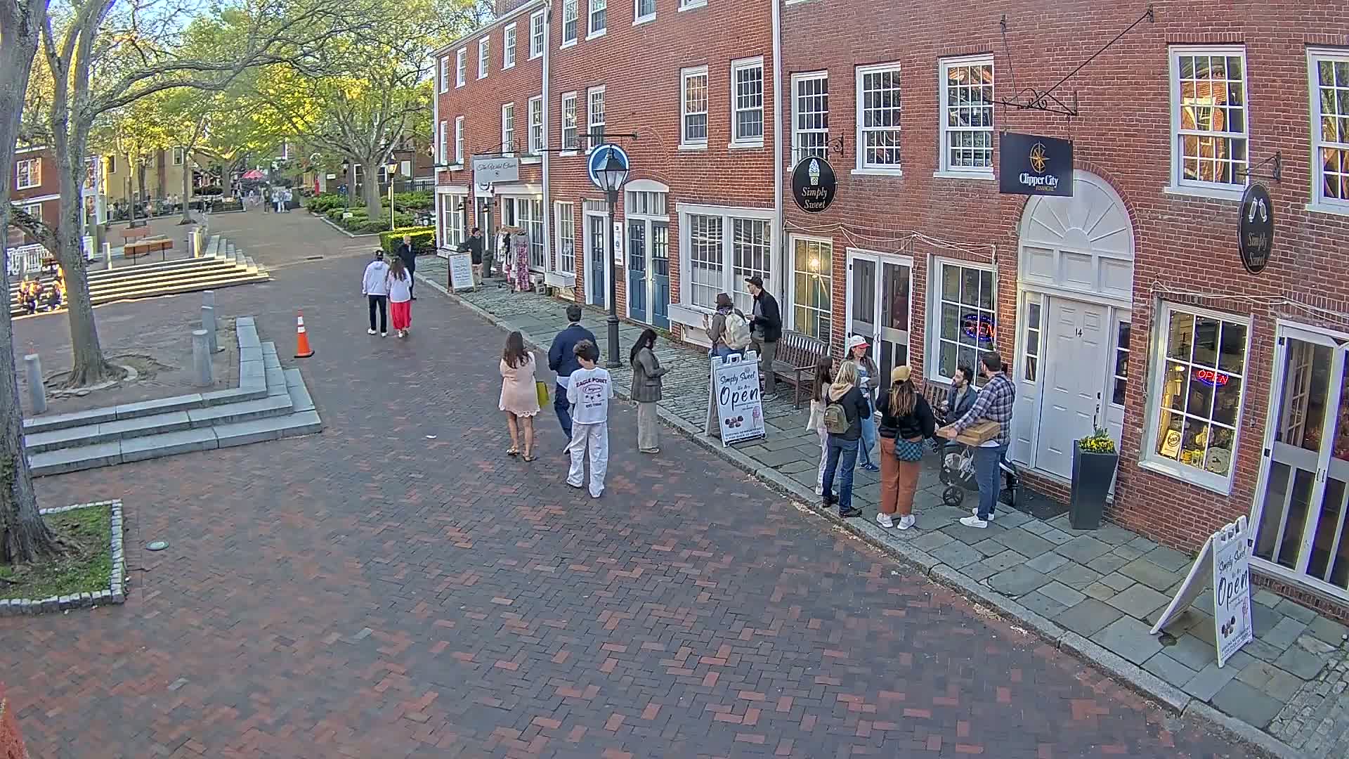 A brick-paved street lined with brick buildings, under a partly sunny sky, shows several people standing in small groups near open storefronts.