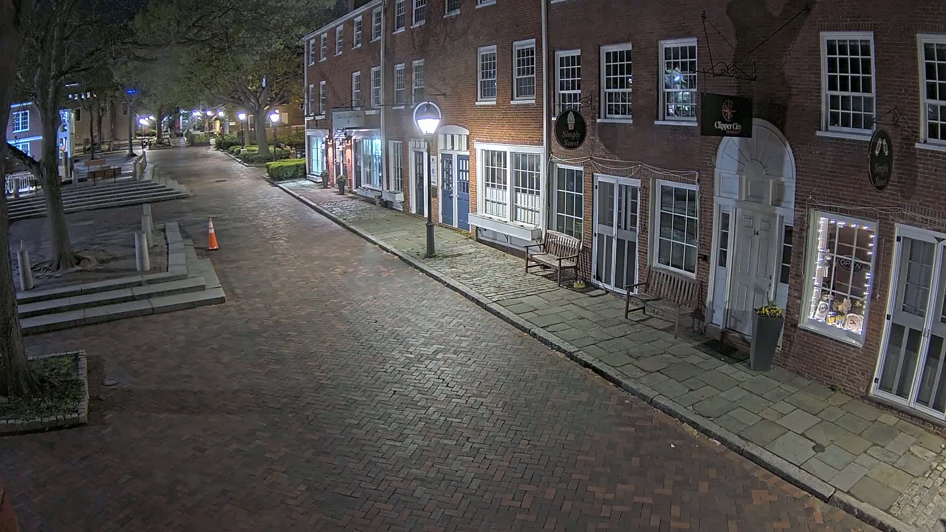 A brick street lined with brick buildings at night, under partly cloudy skies, is illuminated by streetlights.