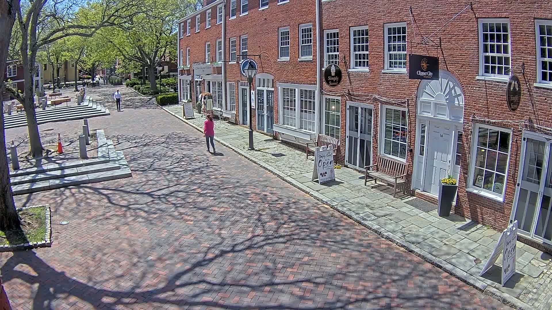 A brick-paved street lined with brick buildings, under a sunny sky with shadows cast by leafy trees, shows two pedestrians walking.