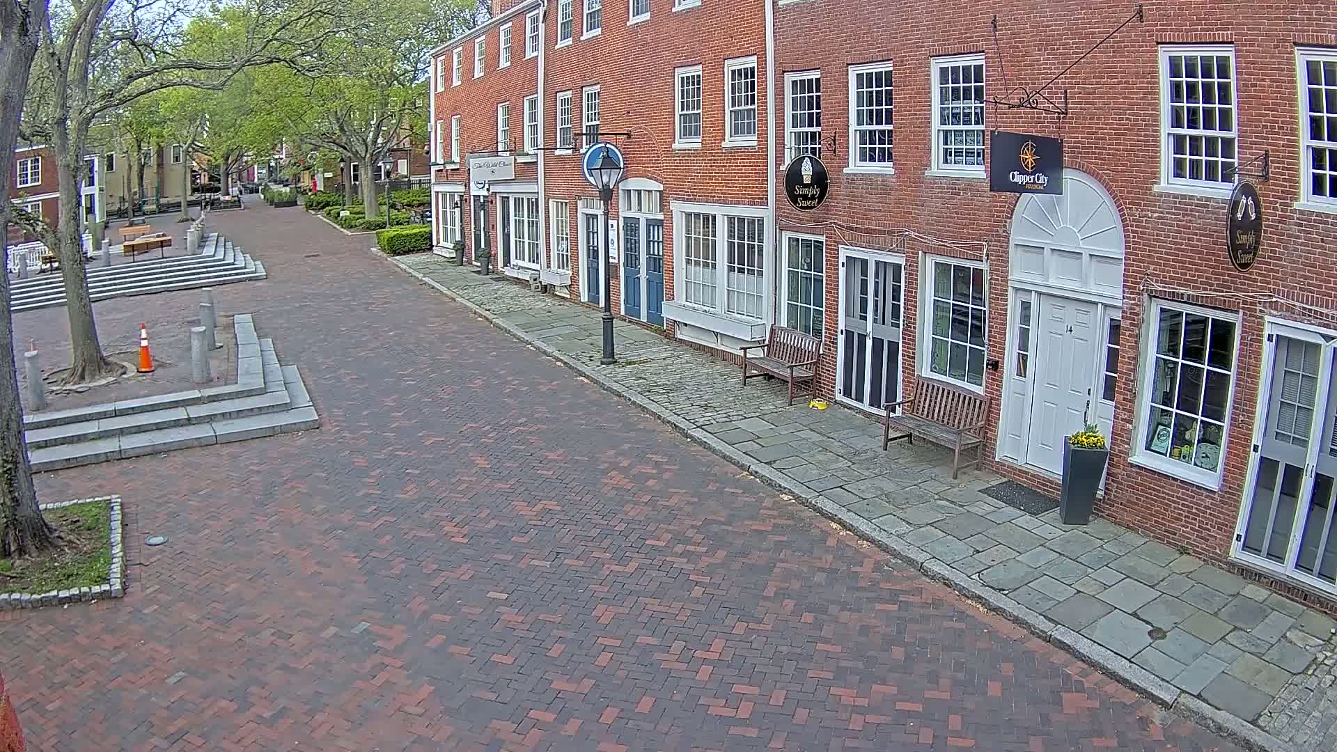 A brick-paved street lined with brick buildings under a partly cloudy sky, showing a quiet, tree-lined town square in the background.