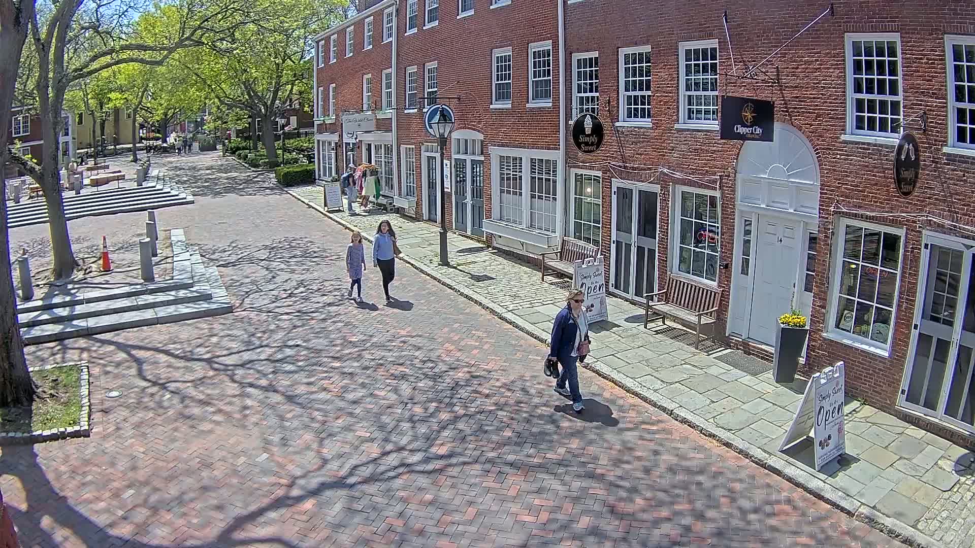 Three people walk down a brick-paved street lined with brick buildings on a sunny day, the shadows of trees falling across the path.