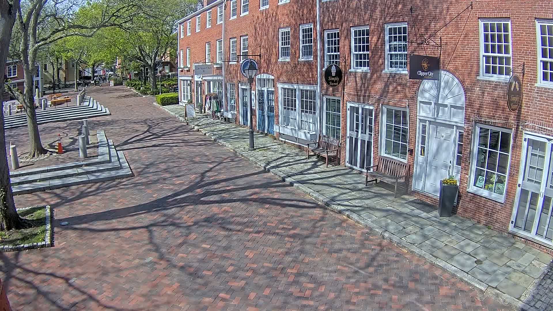 A brick-paved street lined with brick buildings on a sunny day, with tree shadows cast across the pavement.
