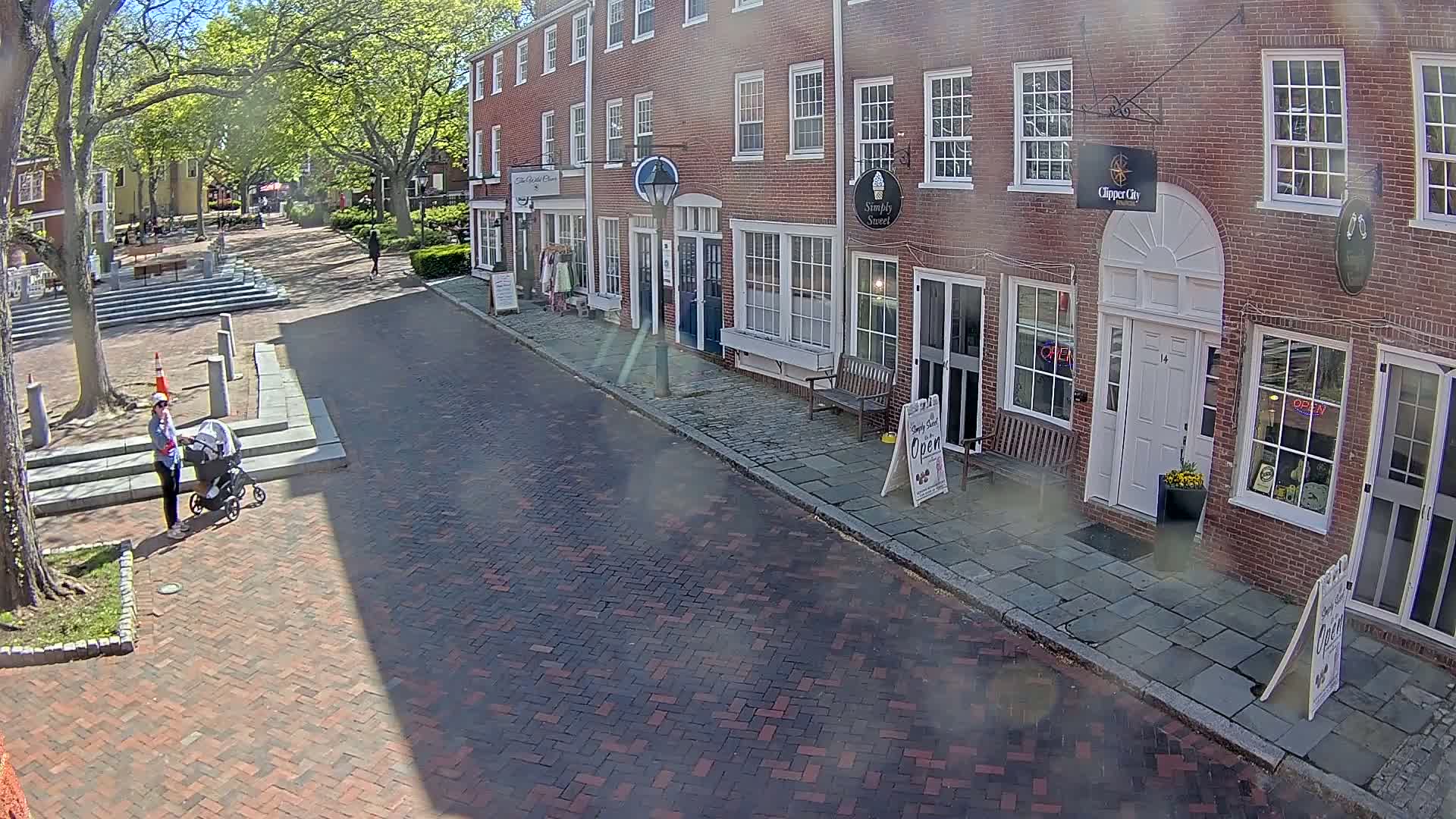 A person pushes a stroller down a brick street lined with brick buildings under a sunny sky.