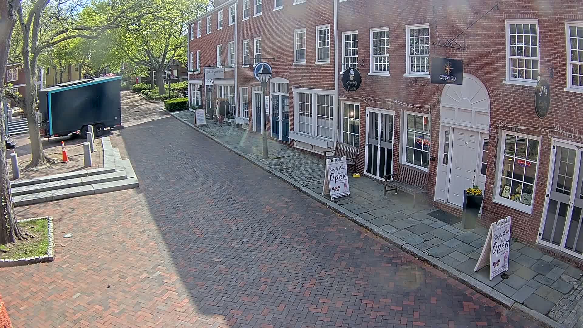 A brick street lined with brick buildings on a sunny day, with a dark-colored trailer parked nearby.