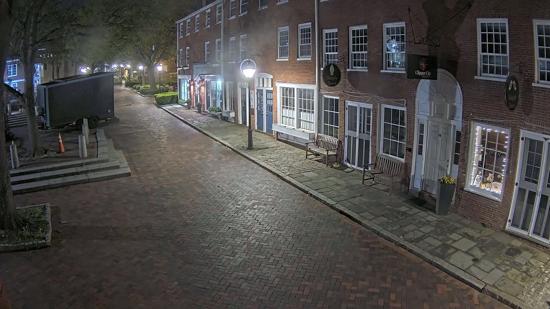A brick-paved street at night, lined with brick buildings and lit by street lamps, shows a mostly empty street with a parked truck at the end.