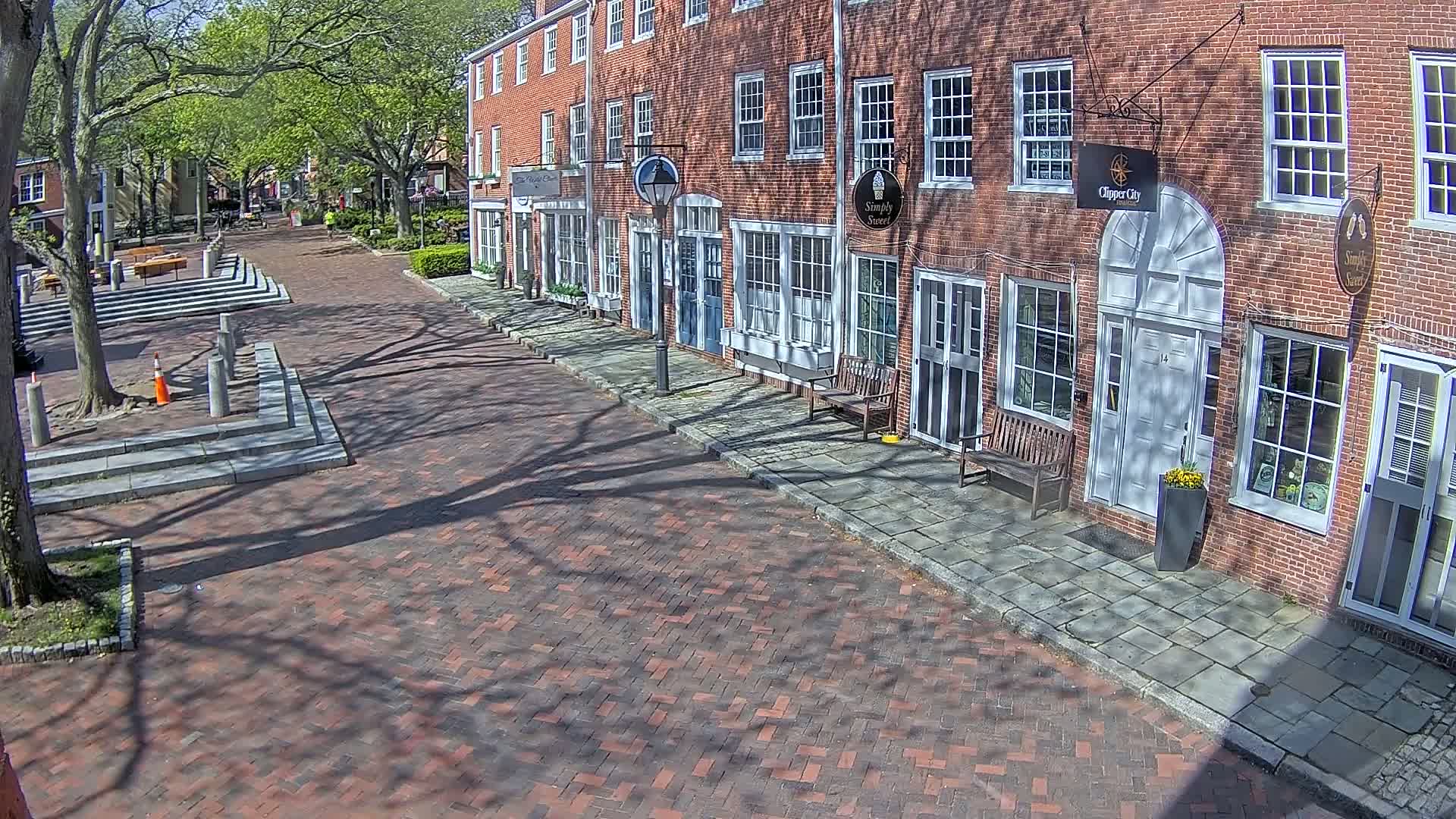 A brick-paved street lined with brick buildings on a sunny day, with the shadows of trees falling across the pavement.