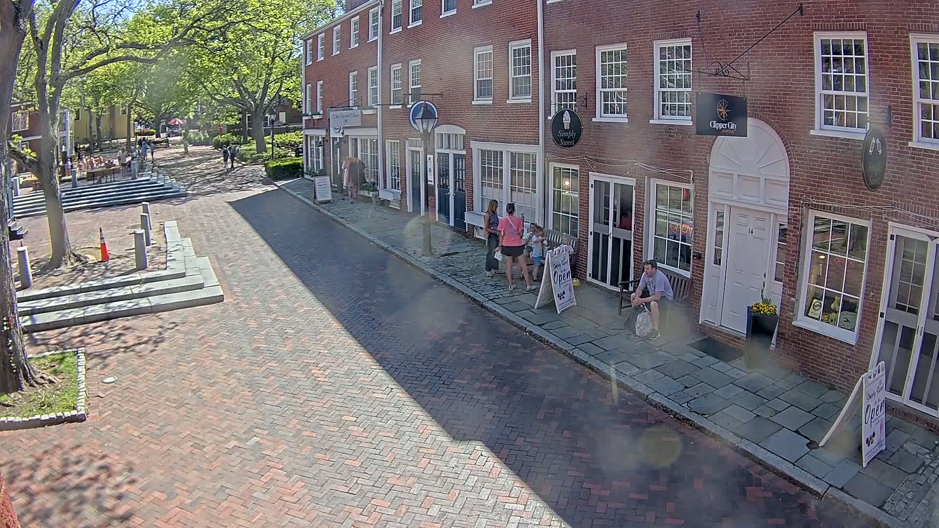 A sunny day on a brick street lined with brick buildings and shops, where several people stand and sit outside.