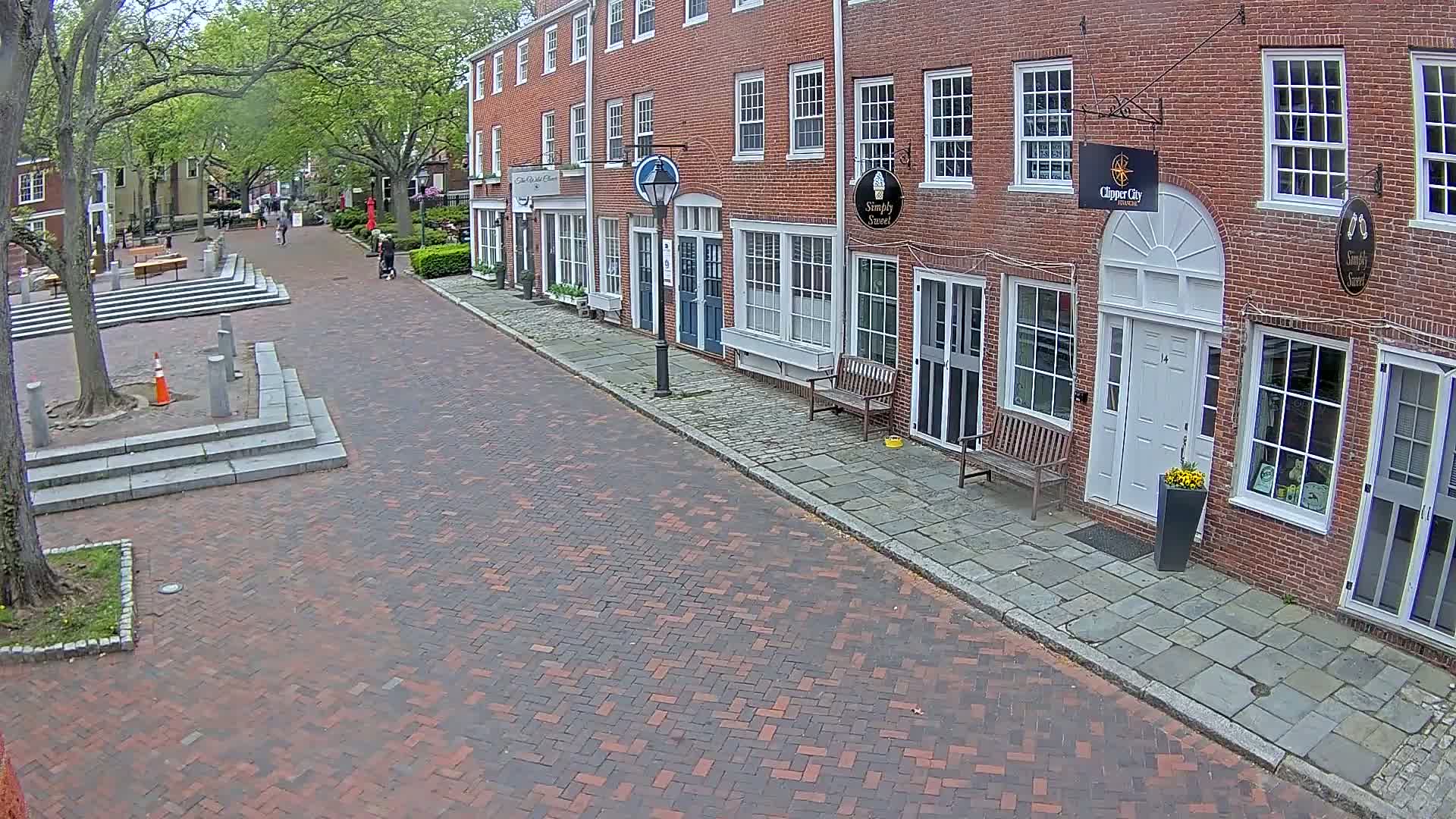 A brick-paved street lined with brick buildings on a partly sunny day, with a few pedestrians visible in the distance.