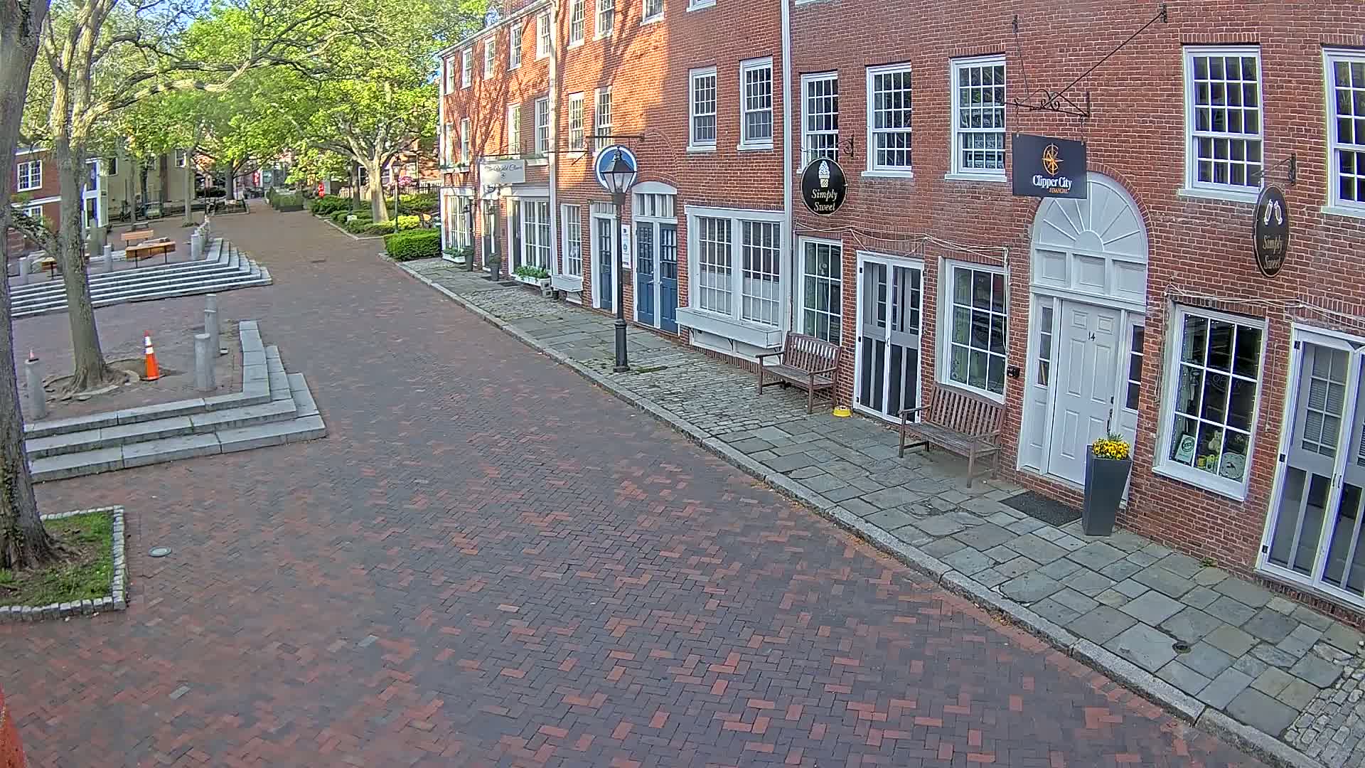 A brick street lined with brick buildings and lush green trees on a sunny day, leading to a brick plaza with tiered seating.