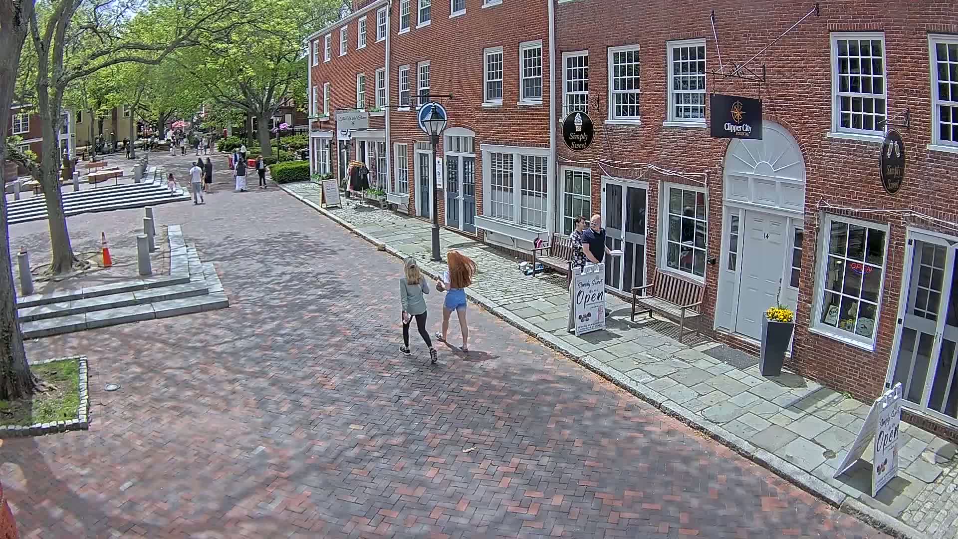 Two women walk down a brick-paved street lined with brick buildings on a sunny day, past a man standing near a shop.