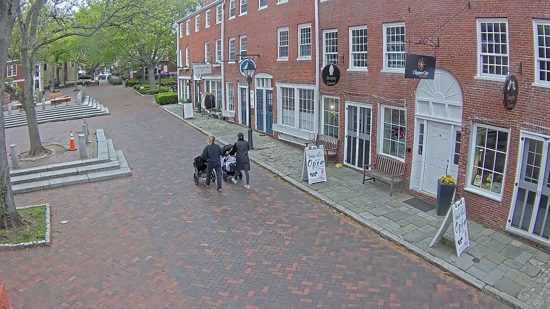 Two adults are pushing strollers down a brick-paved street lined with brick buildings on a partly sunny day.