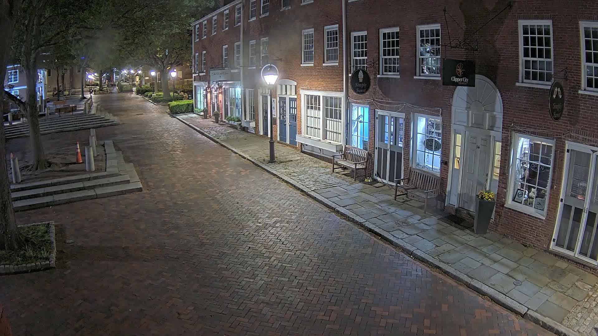 A brick street at night lined with brick buildings containing shops, lit by street lamps under a dark sky.