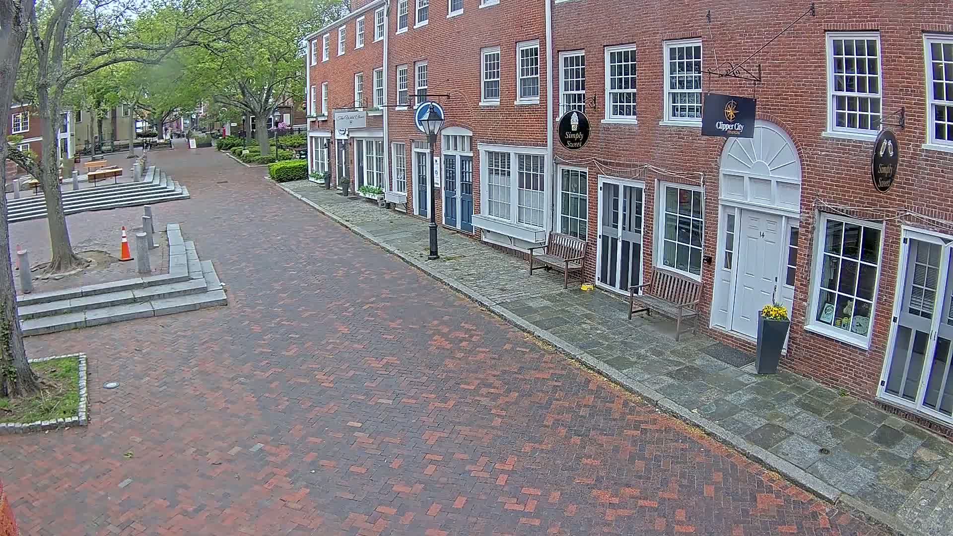 A brick street lined with brick buildings under a partly cloudy sky, featuring a small plaza with stone steps.