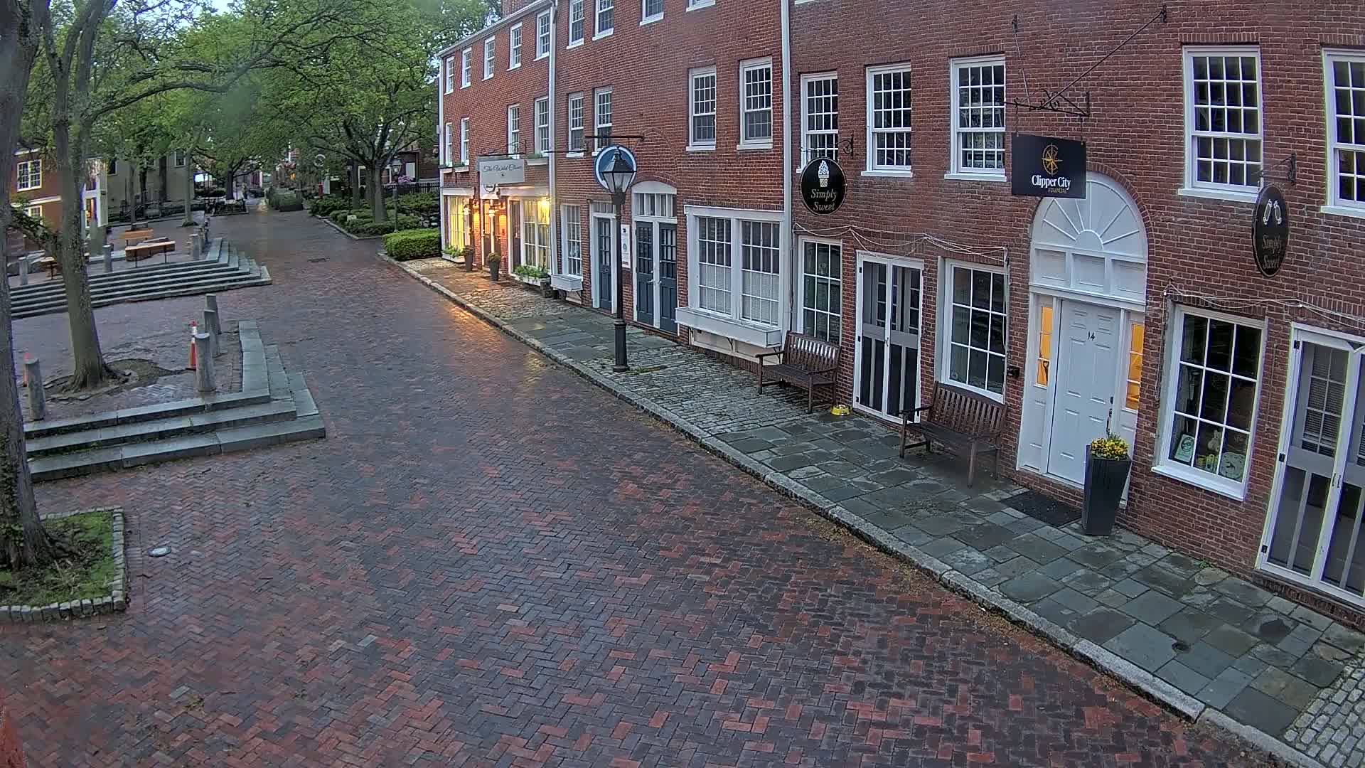A brick street lined with brick buildings under an overcast sky, with some lights on inside the shops.