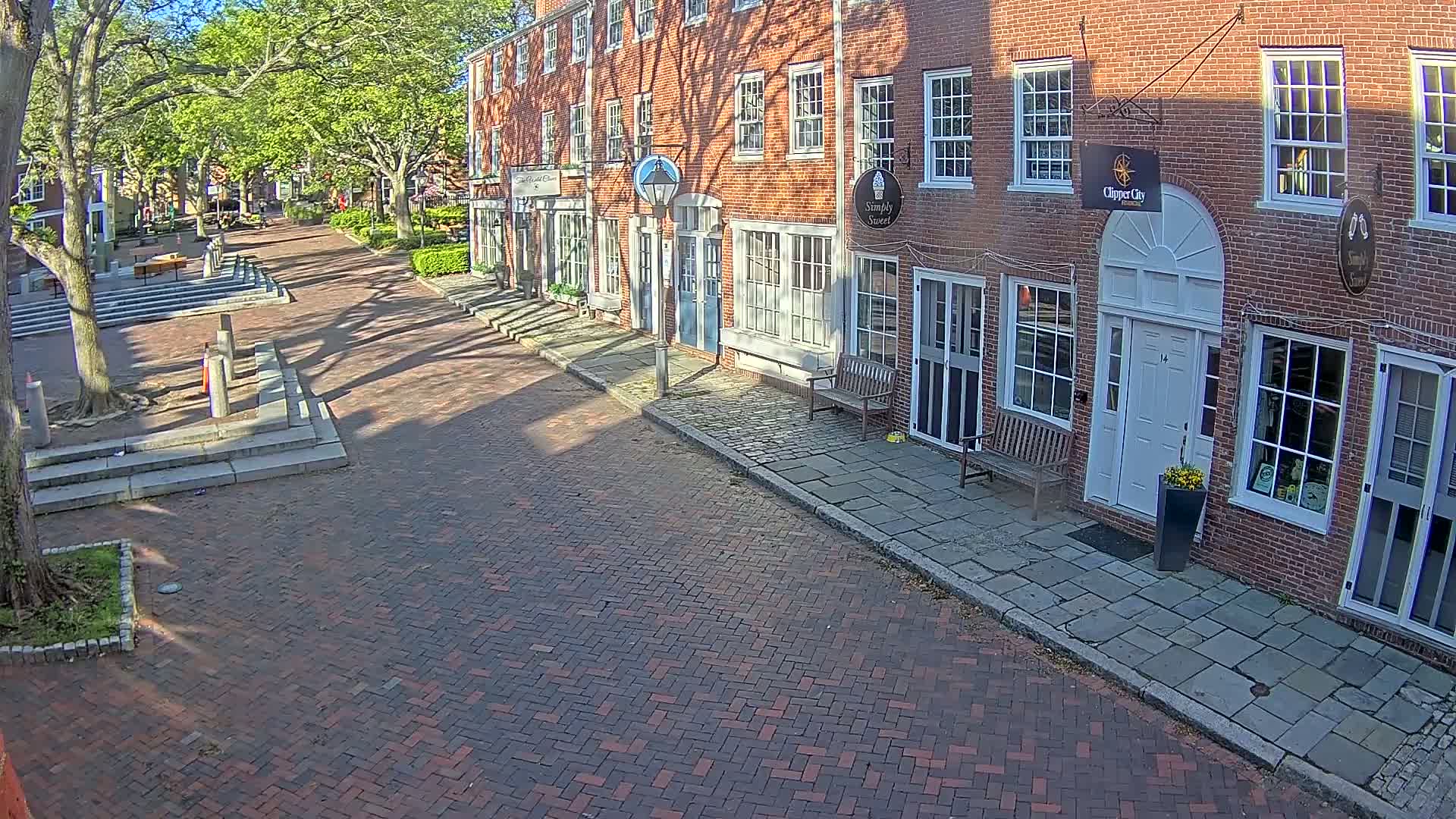 A brick-paved street lined with brick buildings under a sunny sky, with trees providing shade along the sides.
