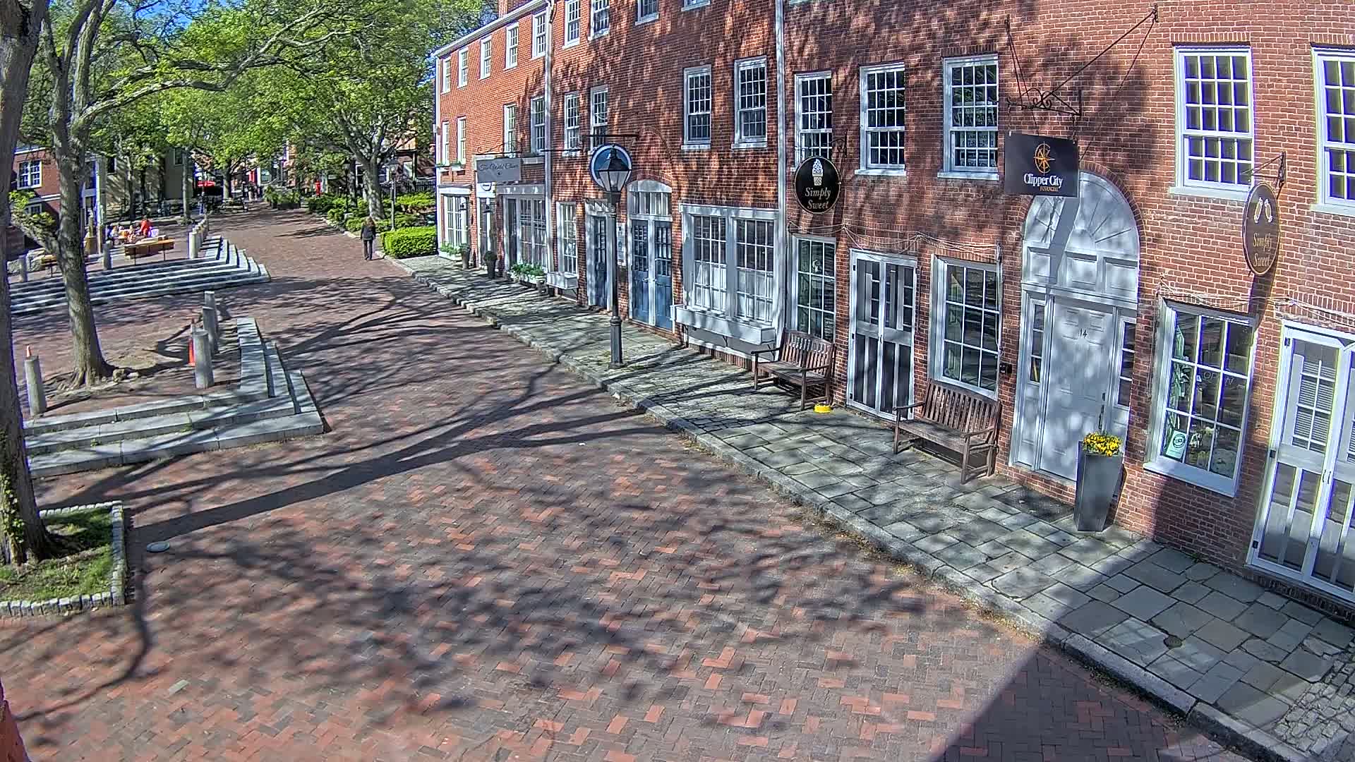 A brick-paved street lined with brick buildings under a sunny sky with tree shadows dappling the ground.