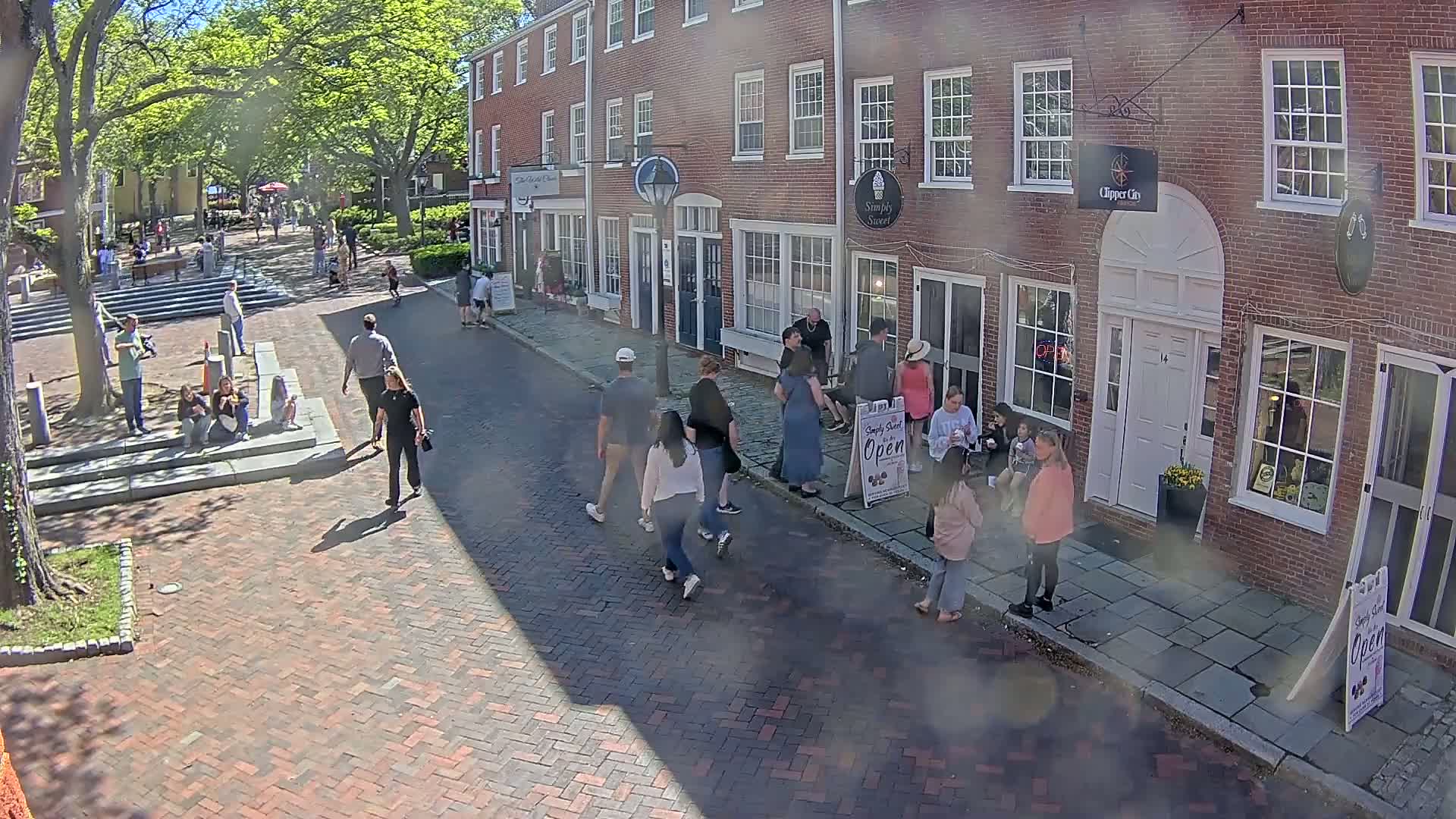 People stroll along a brick-paved street lined with brick buildings on a sunny day, shaded by leafy trees.