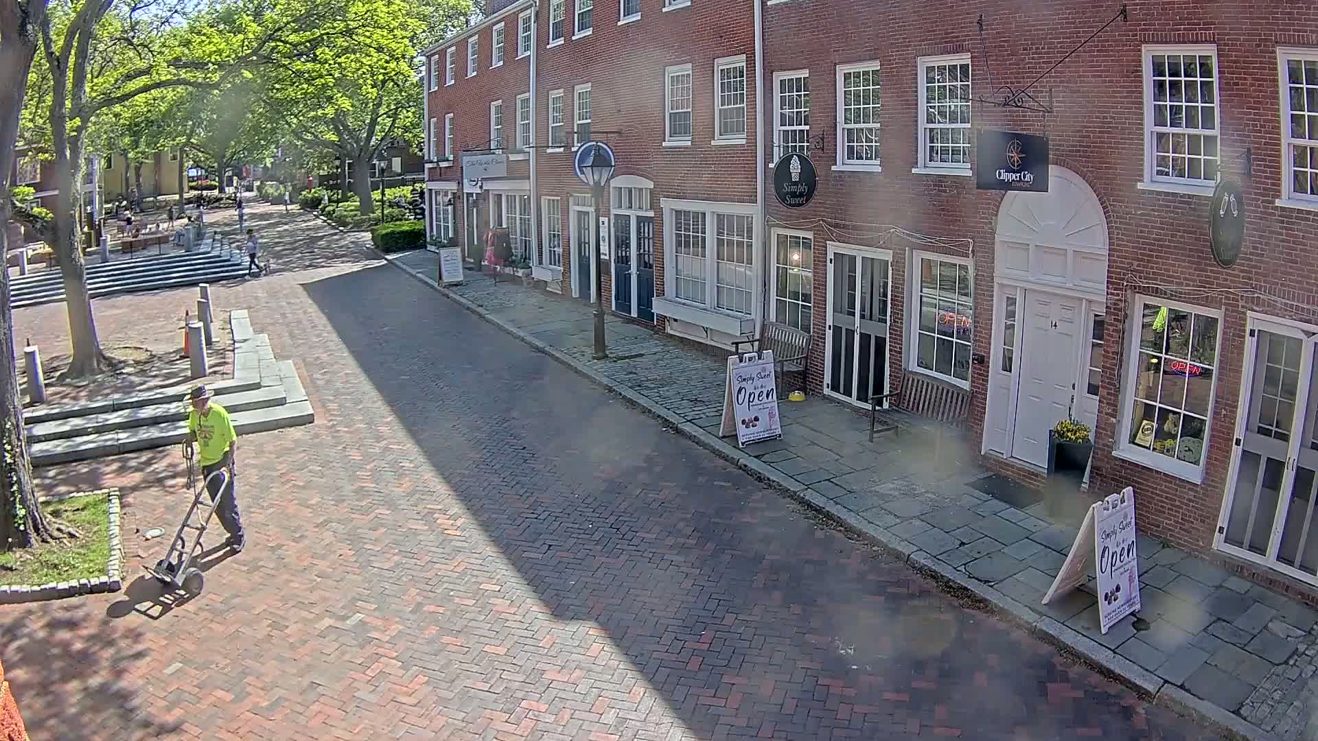 A person in a yellow shirt pushes a cart down a brick-paved street lined with brick buildings on a sunny day.