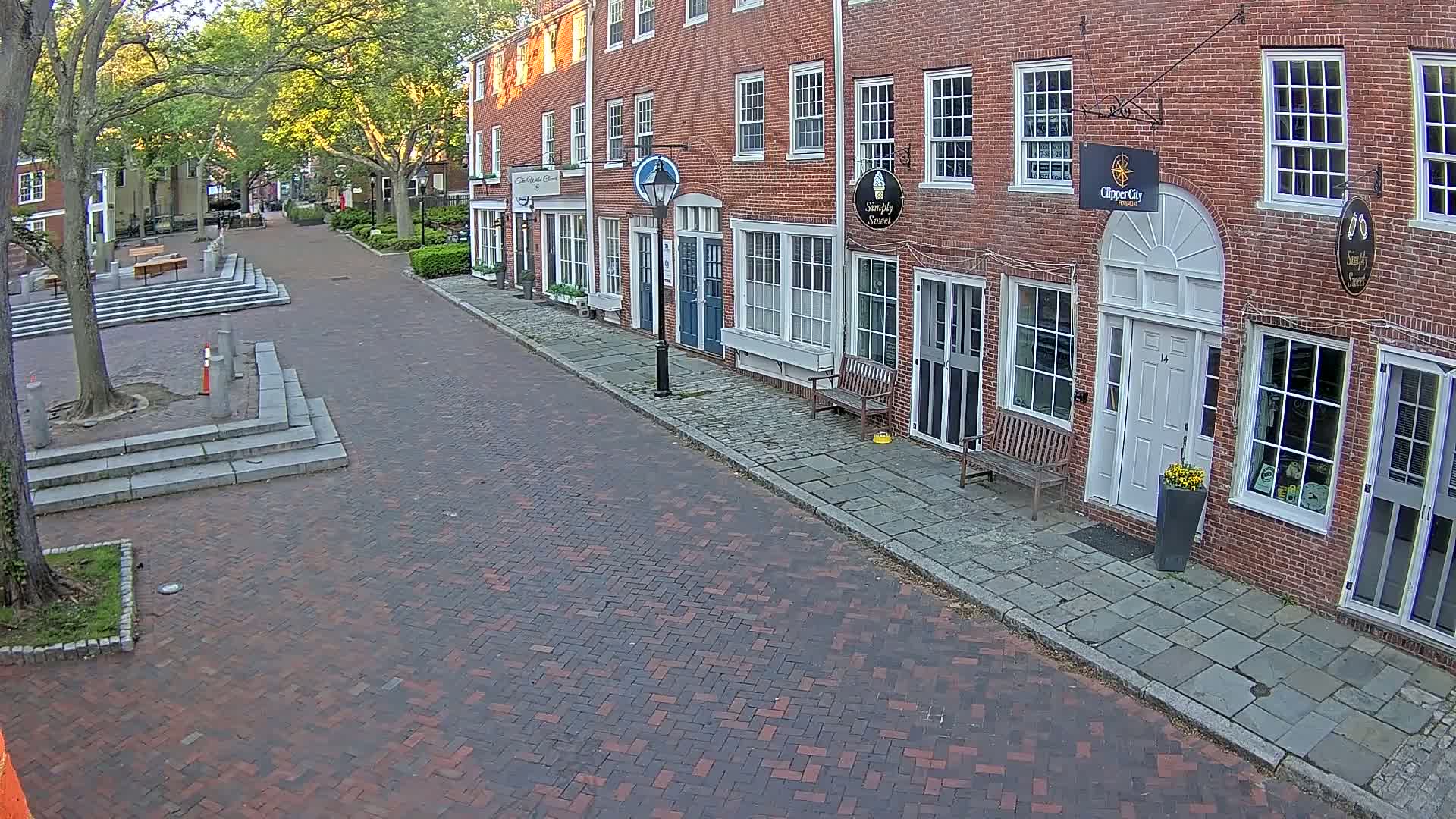 A brick-paved street lined with brick buildings on a sunny day, with a small plaza at the street's beginning.