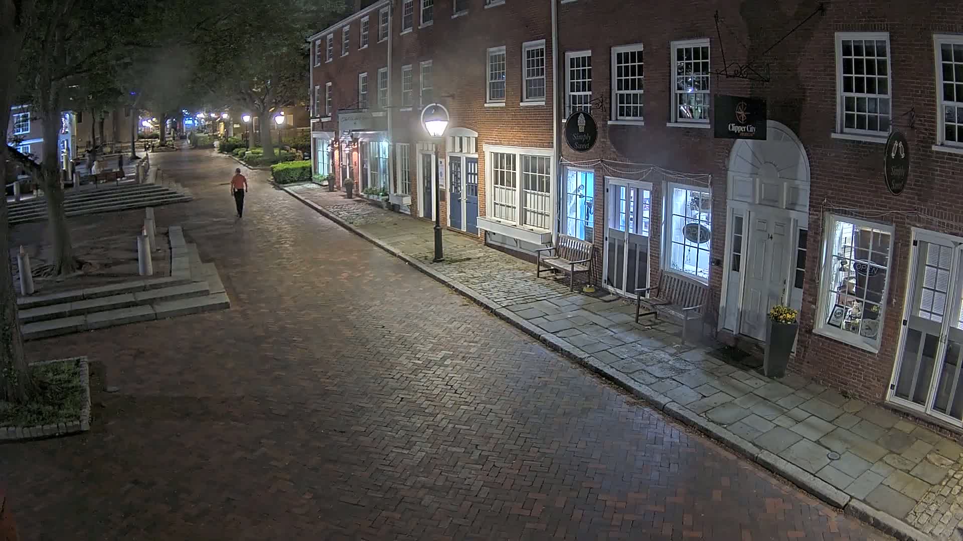 A person walks down a brick-paved street at night past brick buildings with lit shop windows, under a hazy sky.
