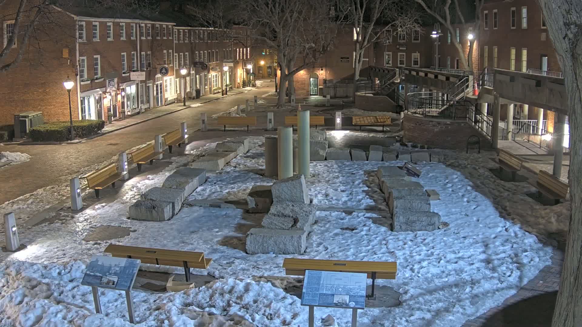 A snow-covered plaza at night, featuring large stone blocks, several benches, and a background of brick buildings.