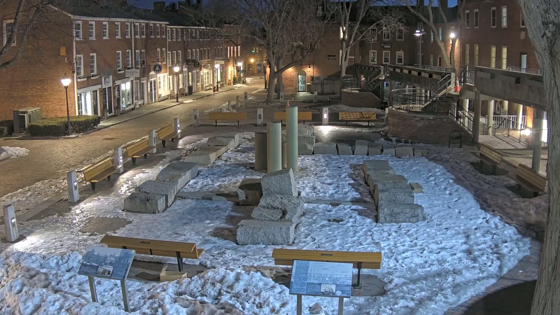 A snow-covered plaza with remnants of stone structures, benches, and streetlights at night, situated between brick buildings.