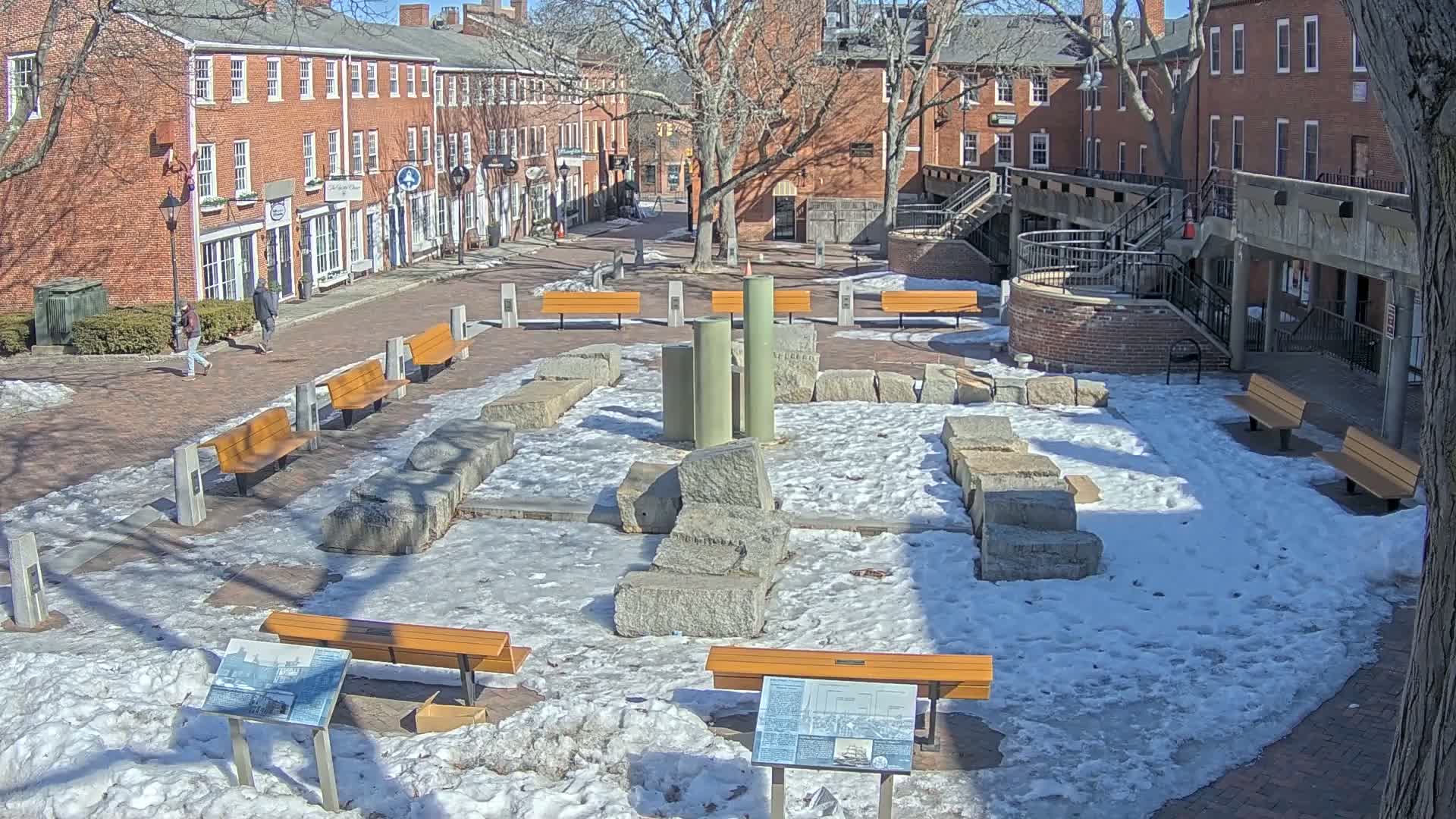 A snow-dusted brick plaza, surrounded by red brick buildings, contains several benches and a central display of large stone blocks and columns.