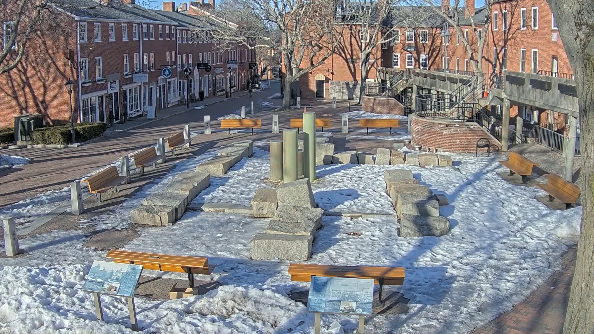 A snow-covered plaza with benches and a stone monument is situated between brick buildings on a sunny day.