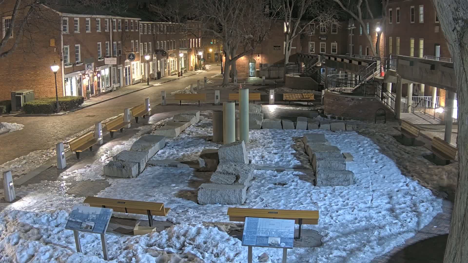 A snow-covered plaza at night, featuring remnants of stone structures, several park benches, and brick buildings lining a street in the background.