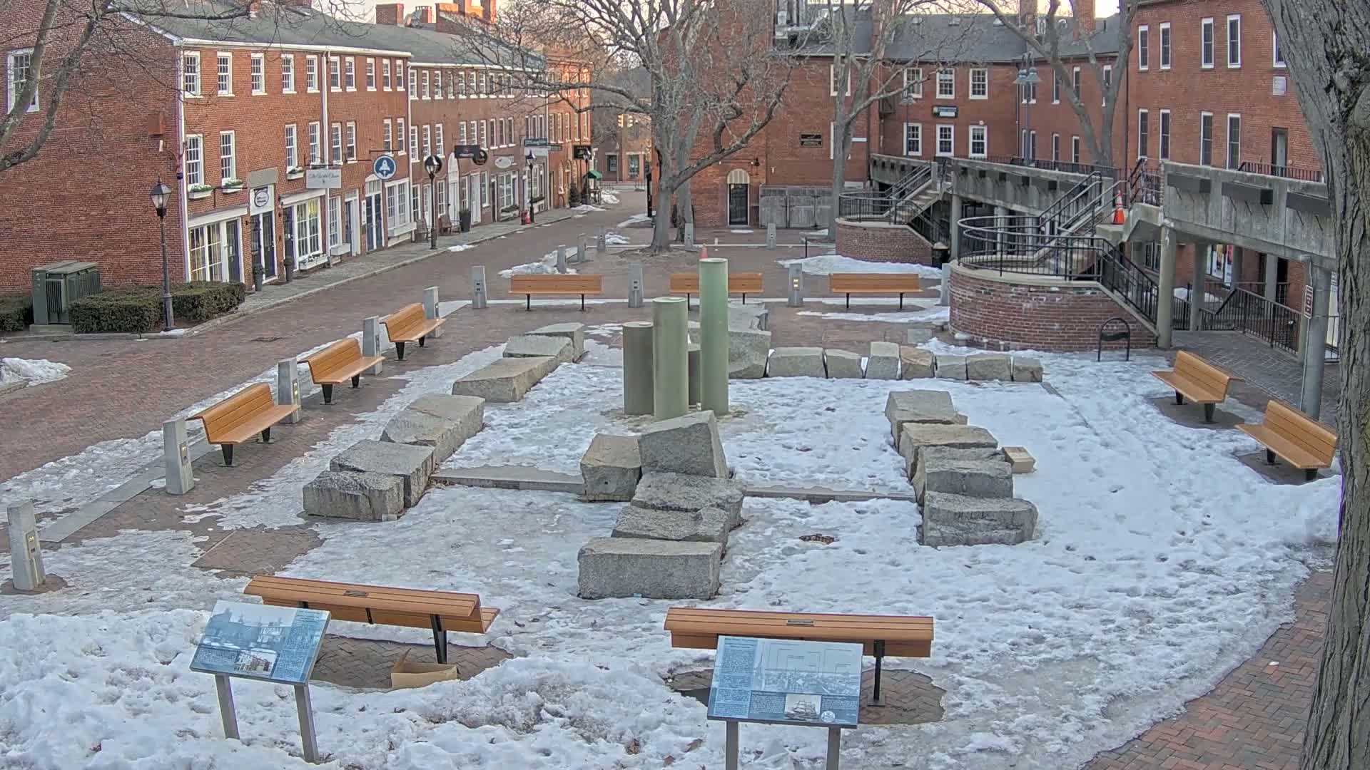 A snow-covered plaza with several benches and a stone arrangement in the center, surrounded by brick buildings.
