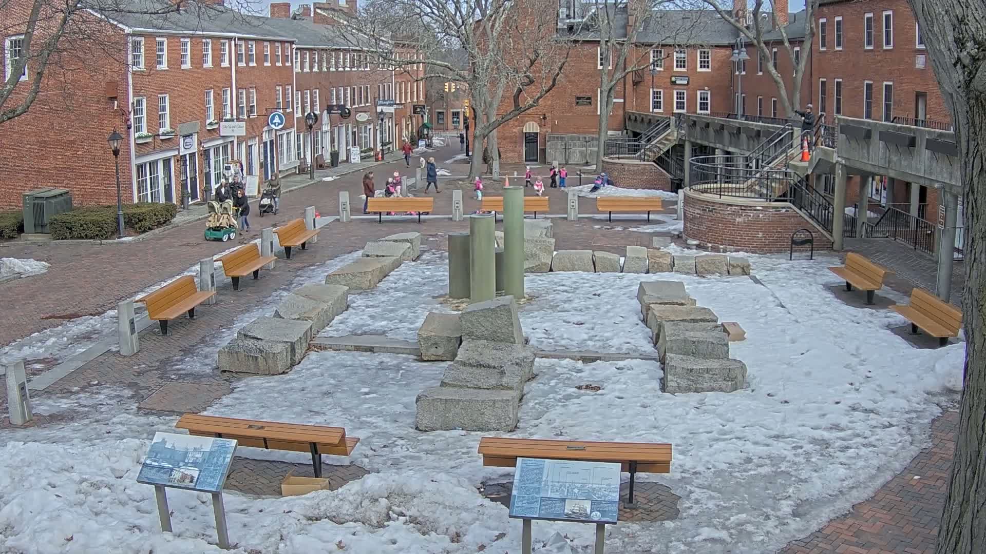 A brick courtyard, partially covered in snow and ice, features a stone monument surrounded by several wooden benches and a few people walking around.