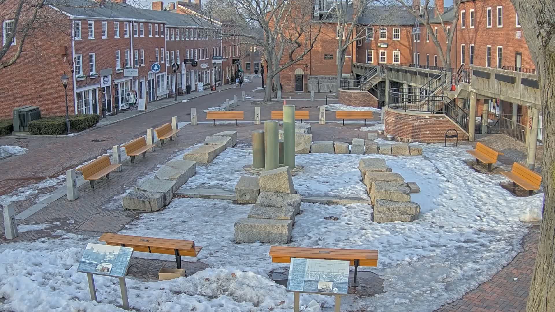 A brick-lined plaza, partially covered in snow, features several wooden benches surrounding a central display of large granite stones and metallic columns.
