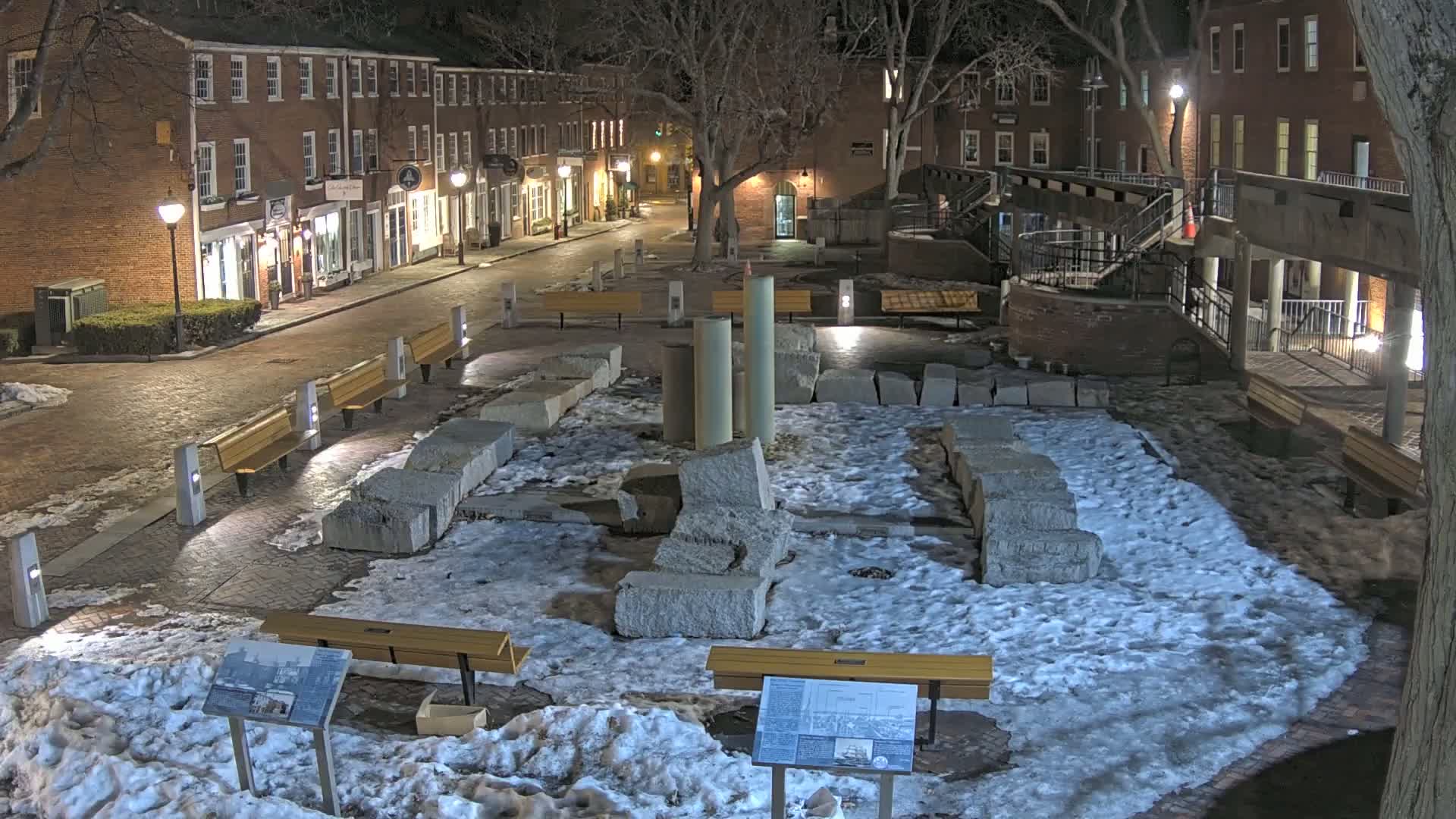 A snow-covered plaza at night, featuring remnants of a stone structure, several benches, and brick buildings lining a street in the background.