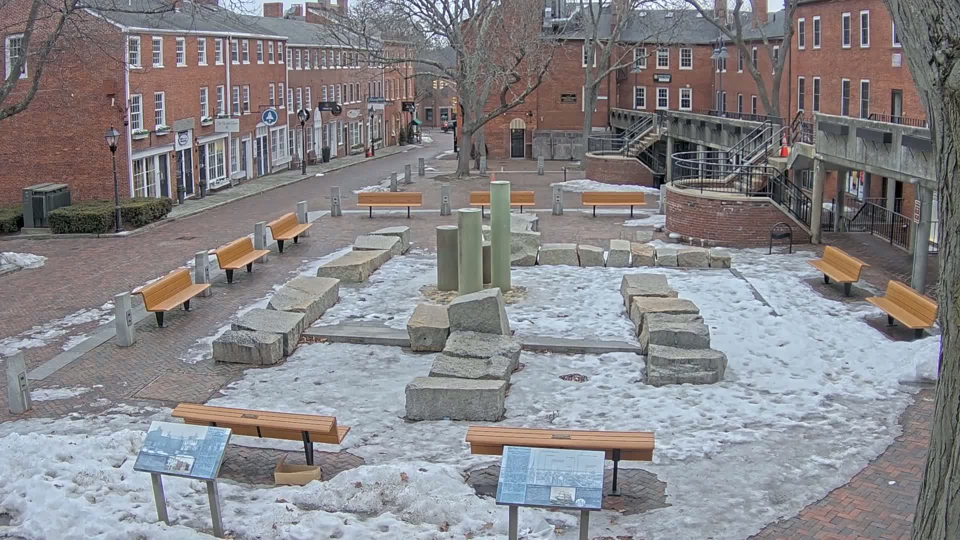 A snow-covered plaza with several benches surrounds a central feature of large granite stones and tall, pale green columns.