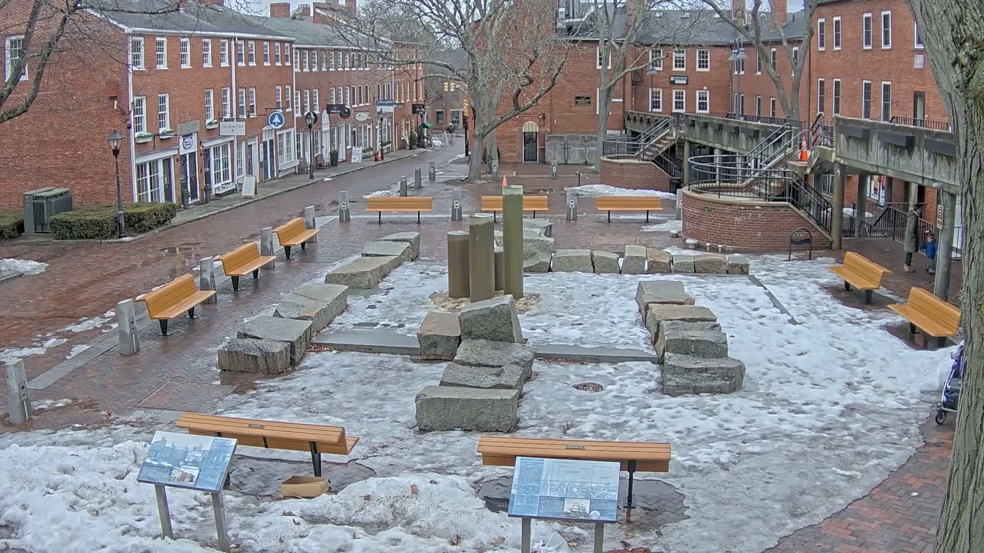 A brick plaza with several benches and a central stone arrangement is partially covered in snow and ice.
