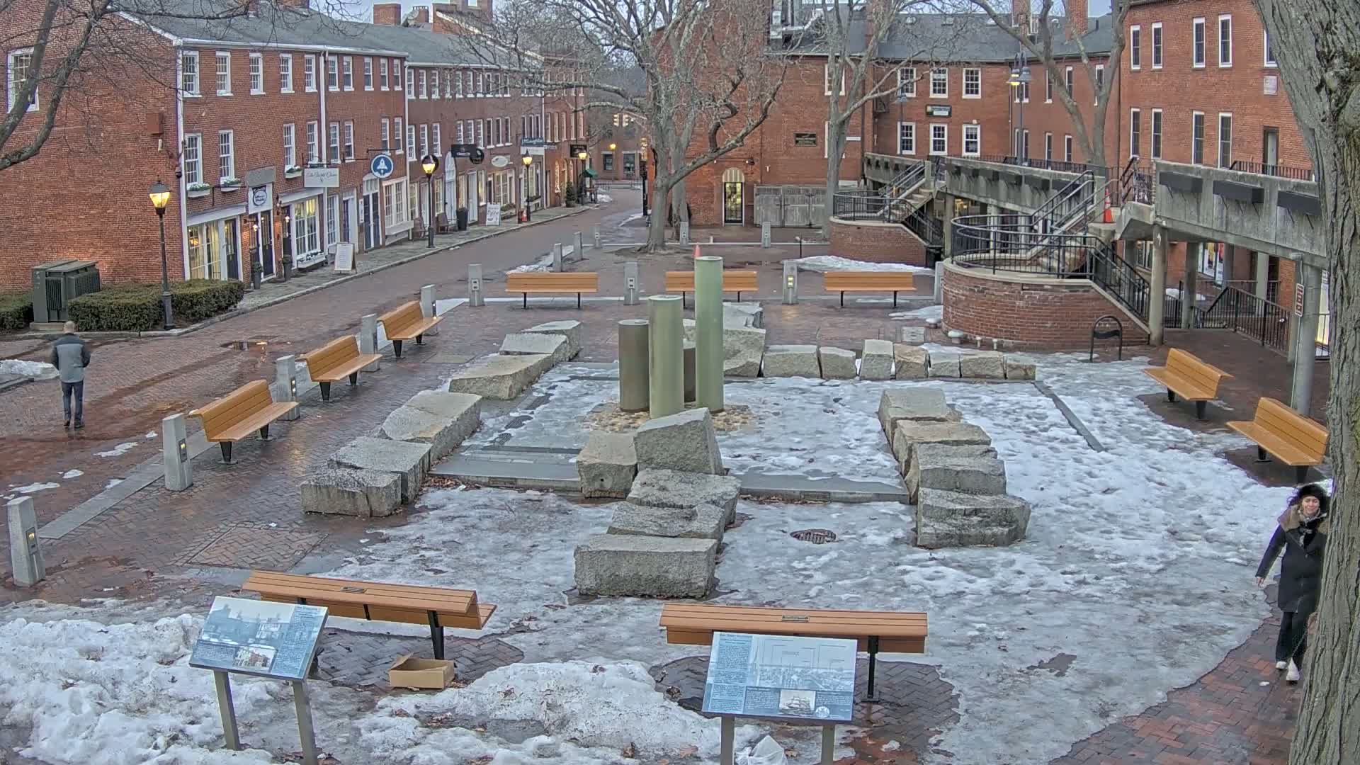 A snow-covered plaza with several benches and a stone fountain is situated between brick buildings on a partly cloudy day.