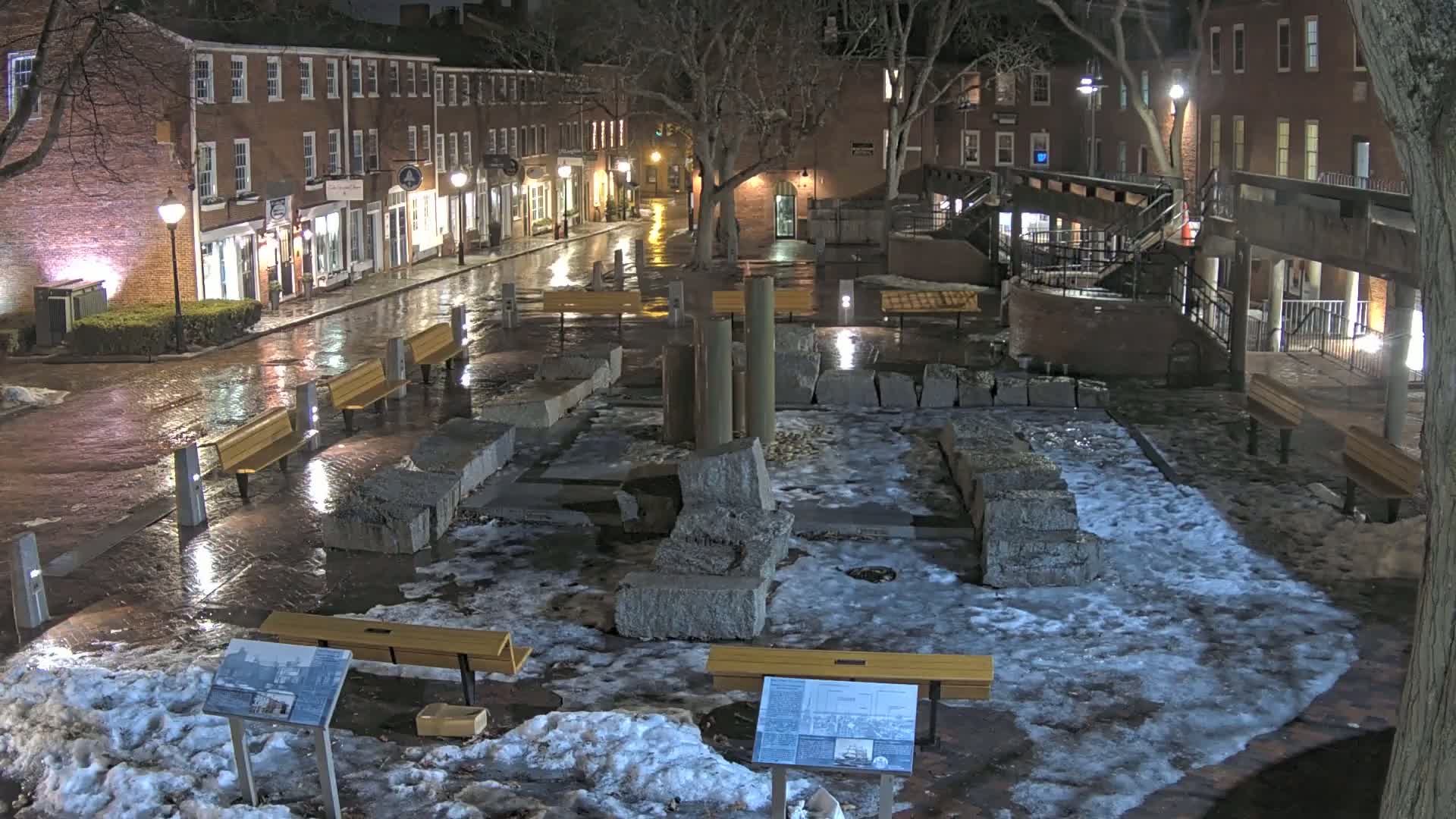 A wet, snow-dusted plaza at night, featuring several benches and a stone structure, is surrounded by brick buildings with streetlights illuminating a rain-slicked street.