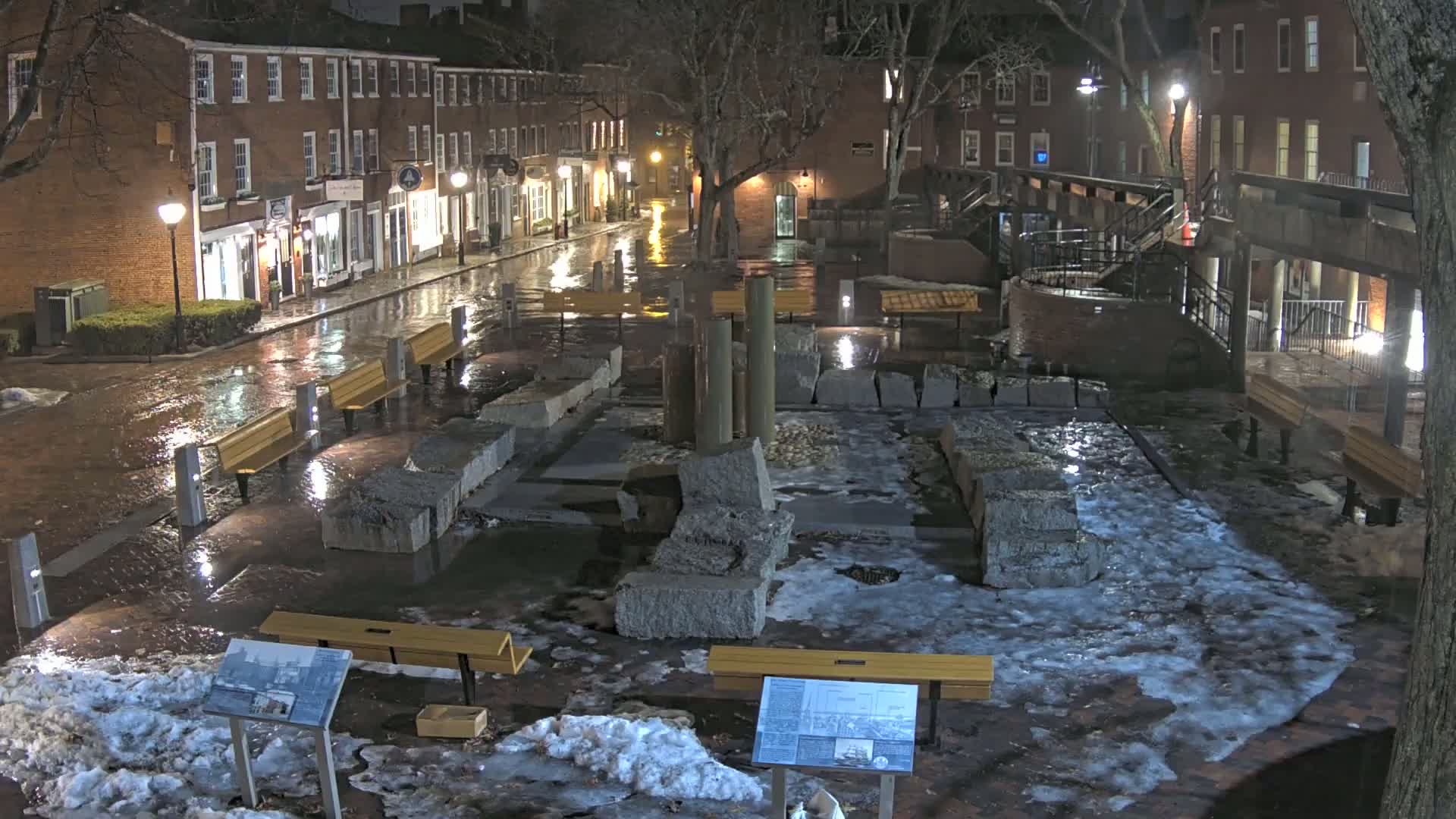 A wet, icy town square at night, featuring stone structures, wooden benches, and brick buildings lining a rain-slicked street.