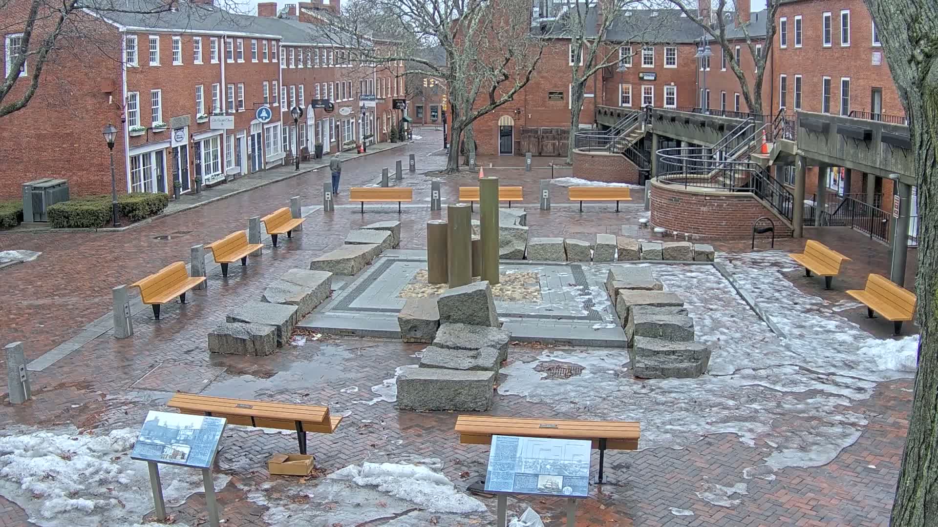 A brick plaza with several wooden benches surrounds a stone fountain partially covered in ice and snow.