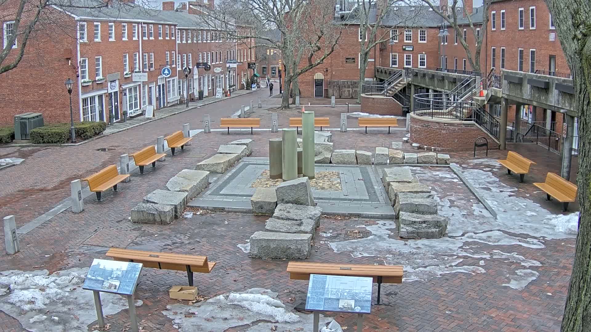 A brick plaza with several benches surrounds a central fountain constructed of large granite stones; patches of snow and ice are present on the ground.
