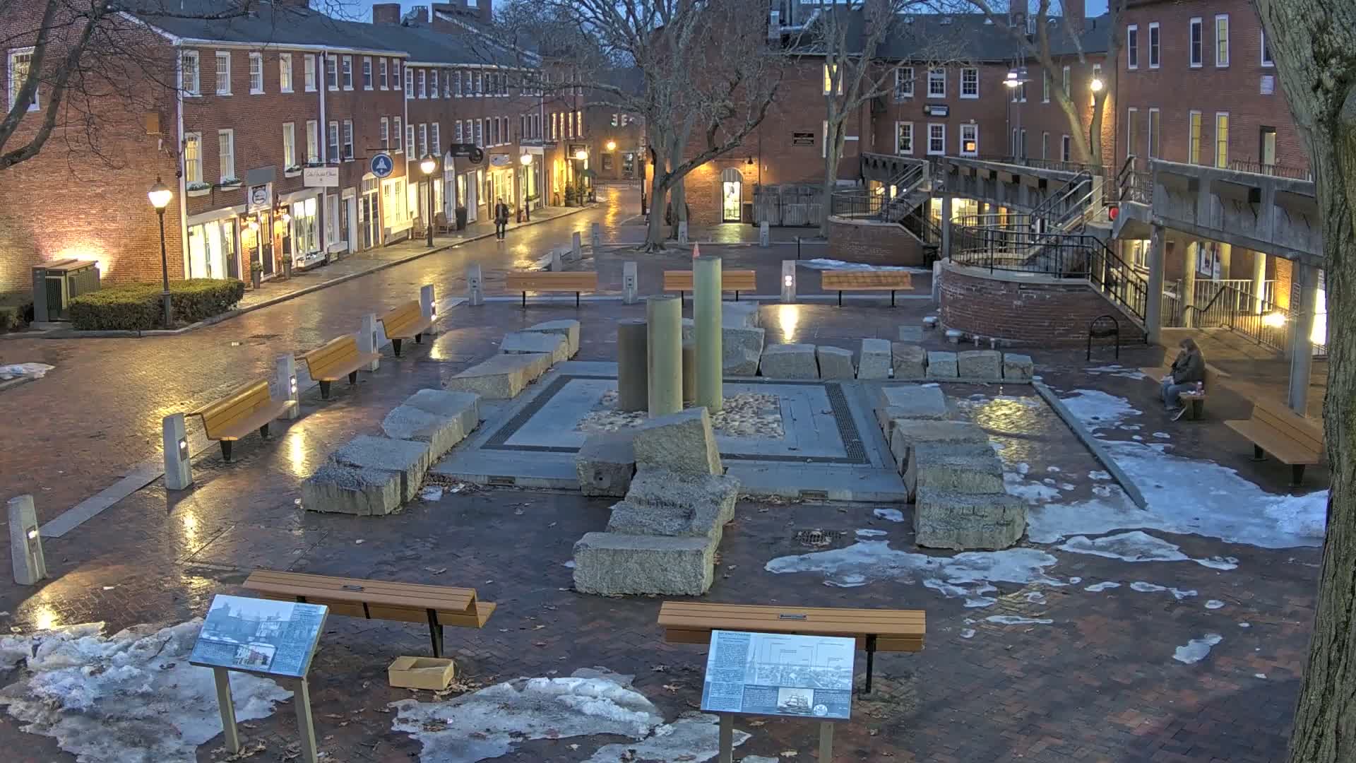 A brick plaza at night with melting snow and ice, featuring a stone monument, several benches, and brick buildings lining the street beyond.