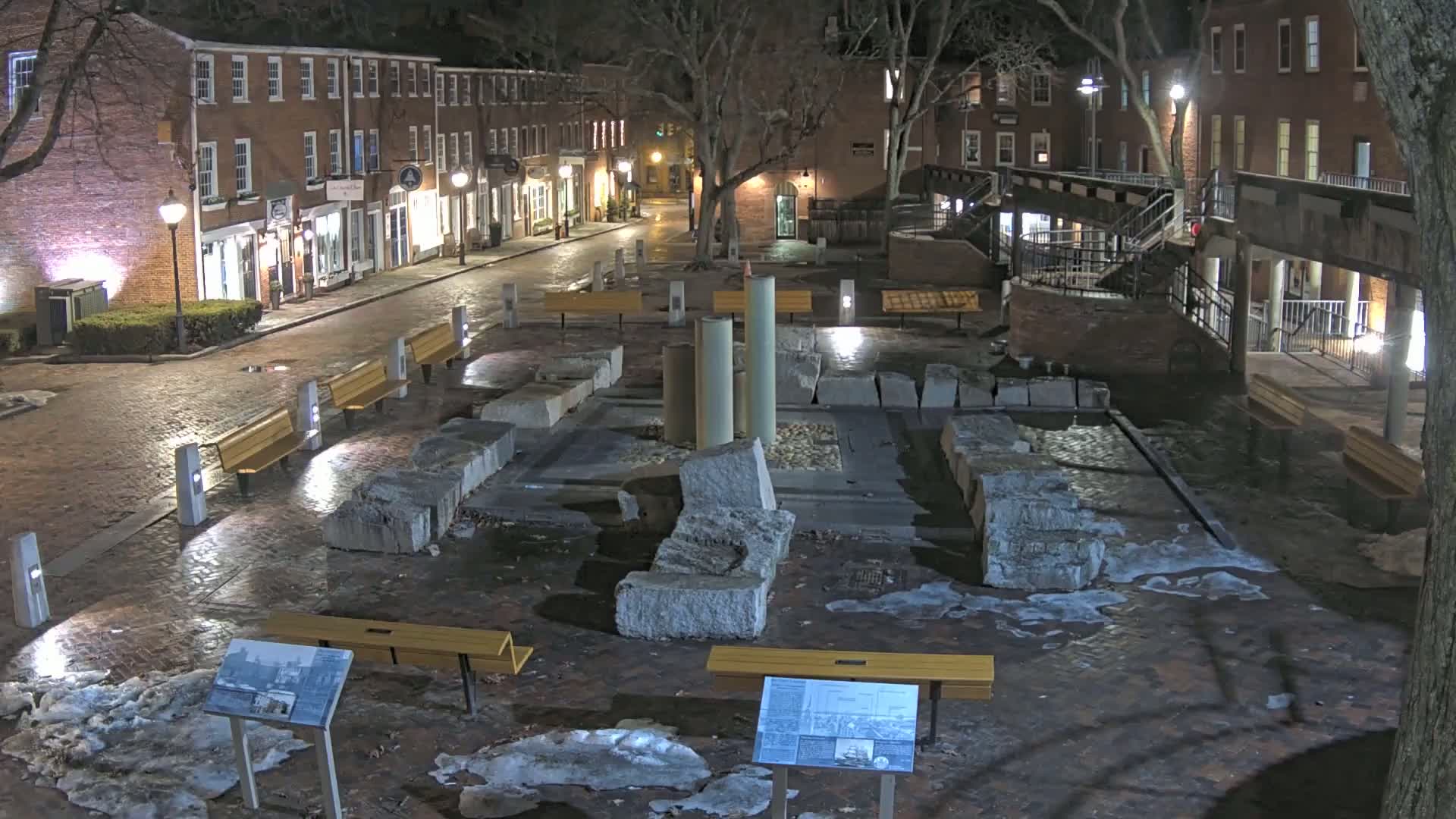 A nighttime view of a brick plaza with a stone fountain, benches, and several buildings lining a street in the background, with patches of snow and ice on the ground.