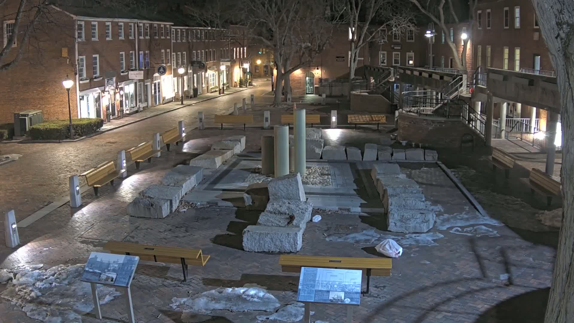 A nighttime view of a brick plaza with a stone monument at its center, surrounded by benches and lit by street lamps, with patches of snow or ice on the ground.