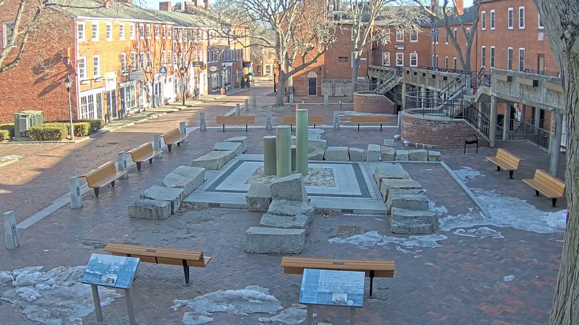 A brick plaza with several benches surrounds a stone and metal fountain, partially covered in snow and ice on a sunny day.